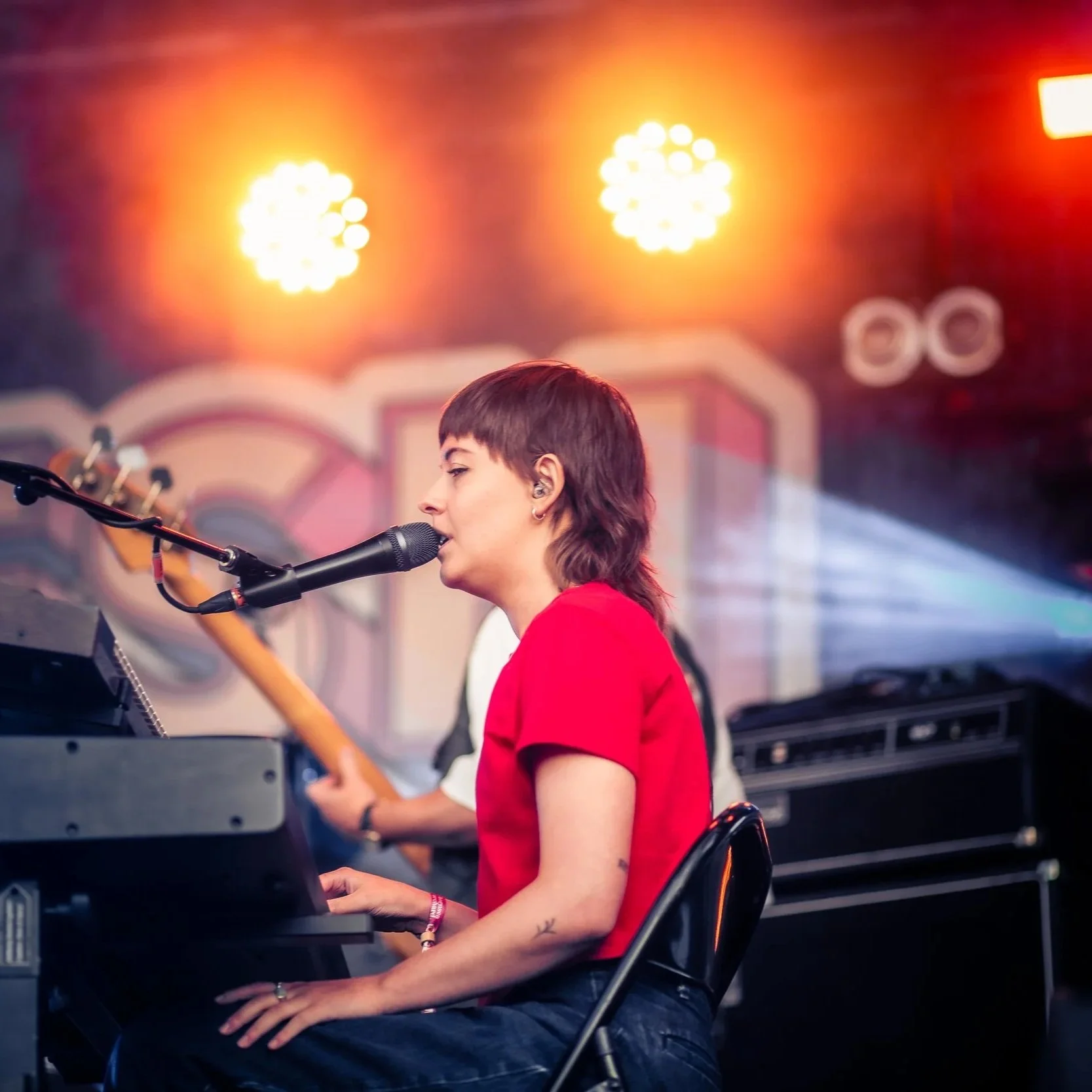 A woman with short dark hair singing into a microphone while playing an electronic keyboard on stage, with a guitar player behind her and colorful stage lights above.