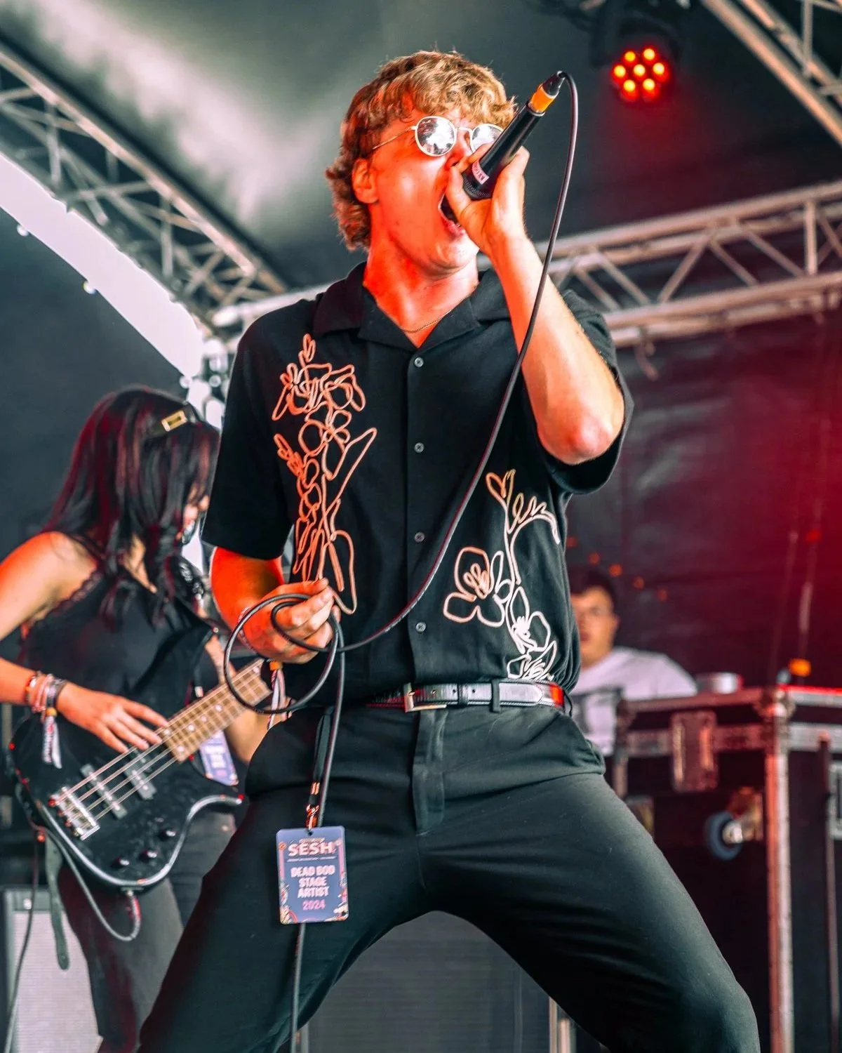 A man singing into a microphone on stage at a concert, wearing round sunglasses and a black shirt with floral embroidery. There is a woman in the background playing a bass guitar.