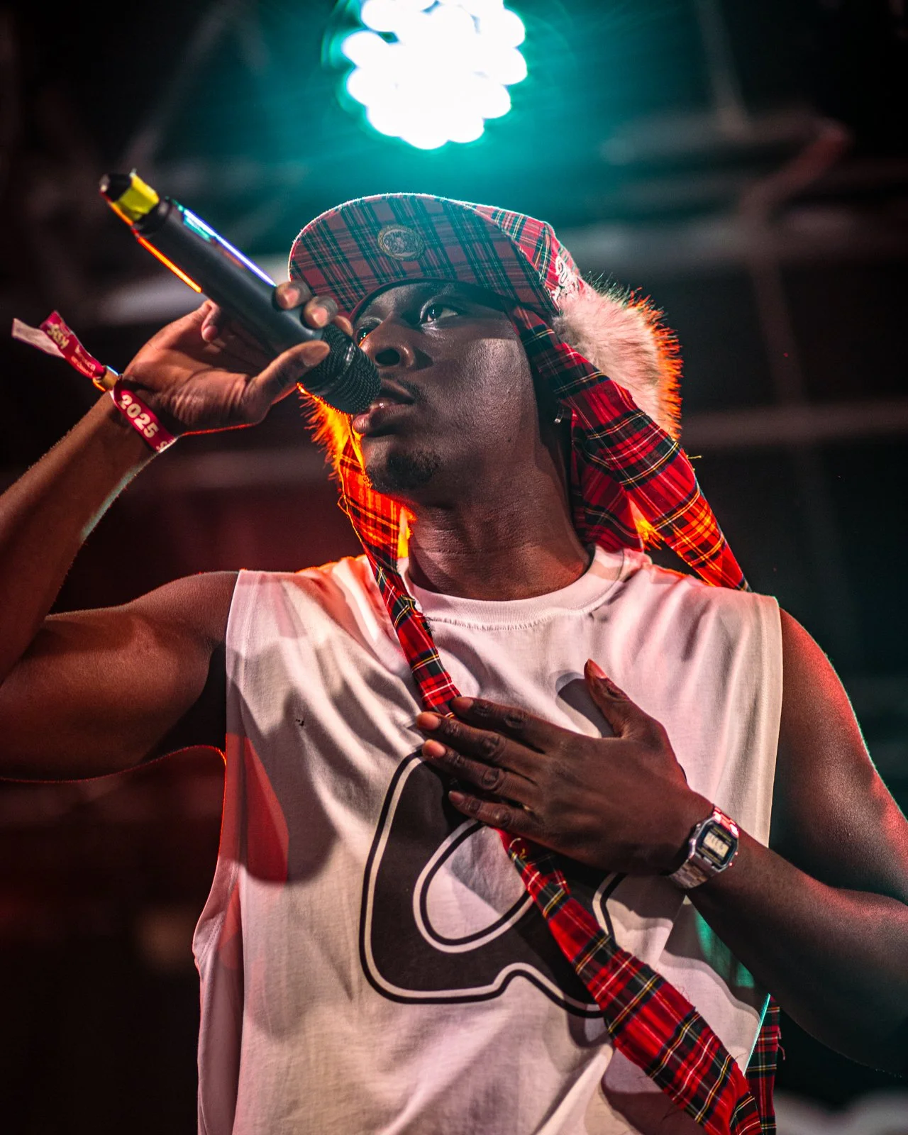 A man singing into a microphone on stage, wearing a patterned headscarf, sleeveless shirt, and a wristwatch, with one hand on his chest and a colorful light above.