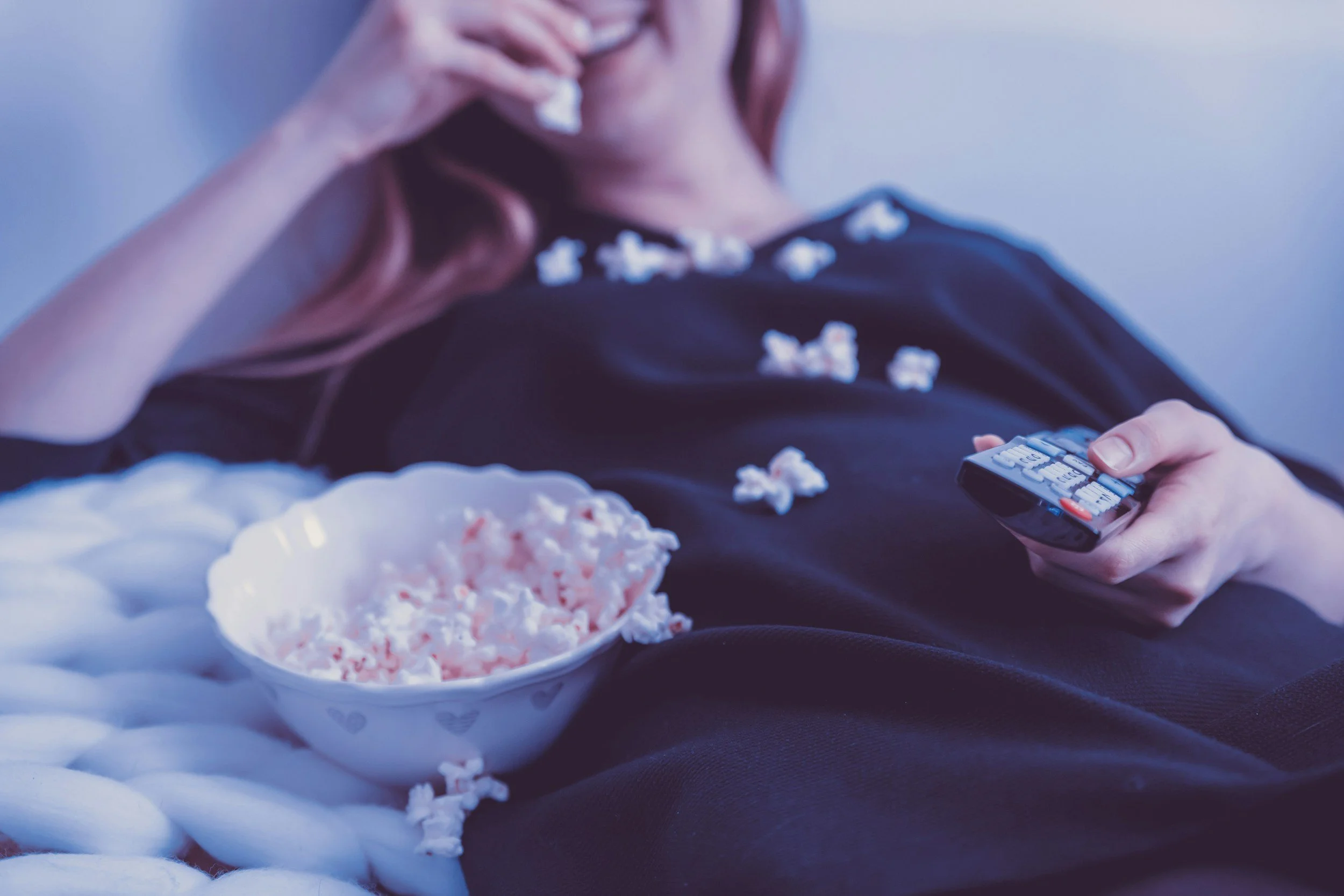 Person in black clothing lying on a bed, holding a remote control in one hand and eating popcorn with the other, with an empty bowl of popcorn nearby.