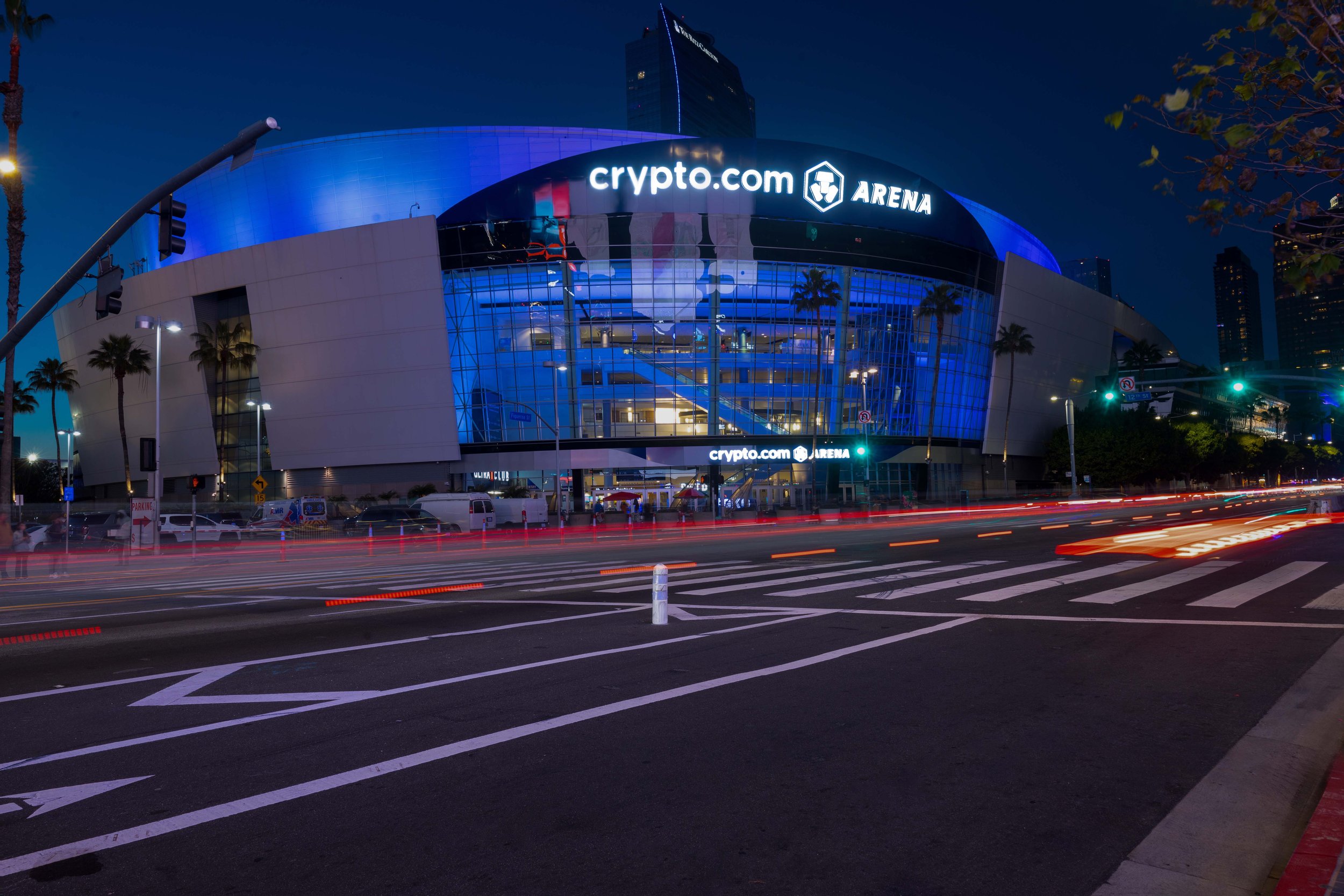 Nighttime exterior view of the crypto.com arena, a large modern building with blue lighting and glass facade, displaying the crypto.com logo and the word arena, with light streaks from passing cars in the foreground.