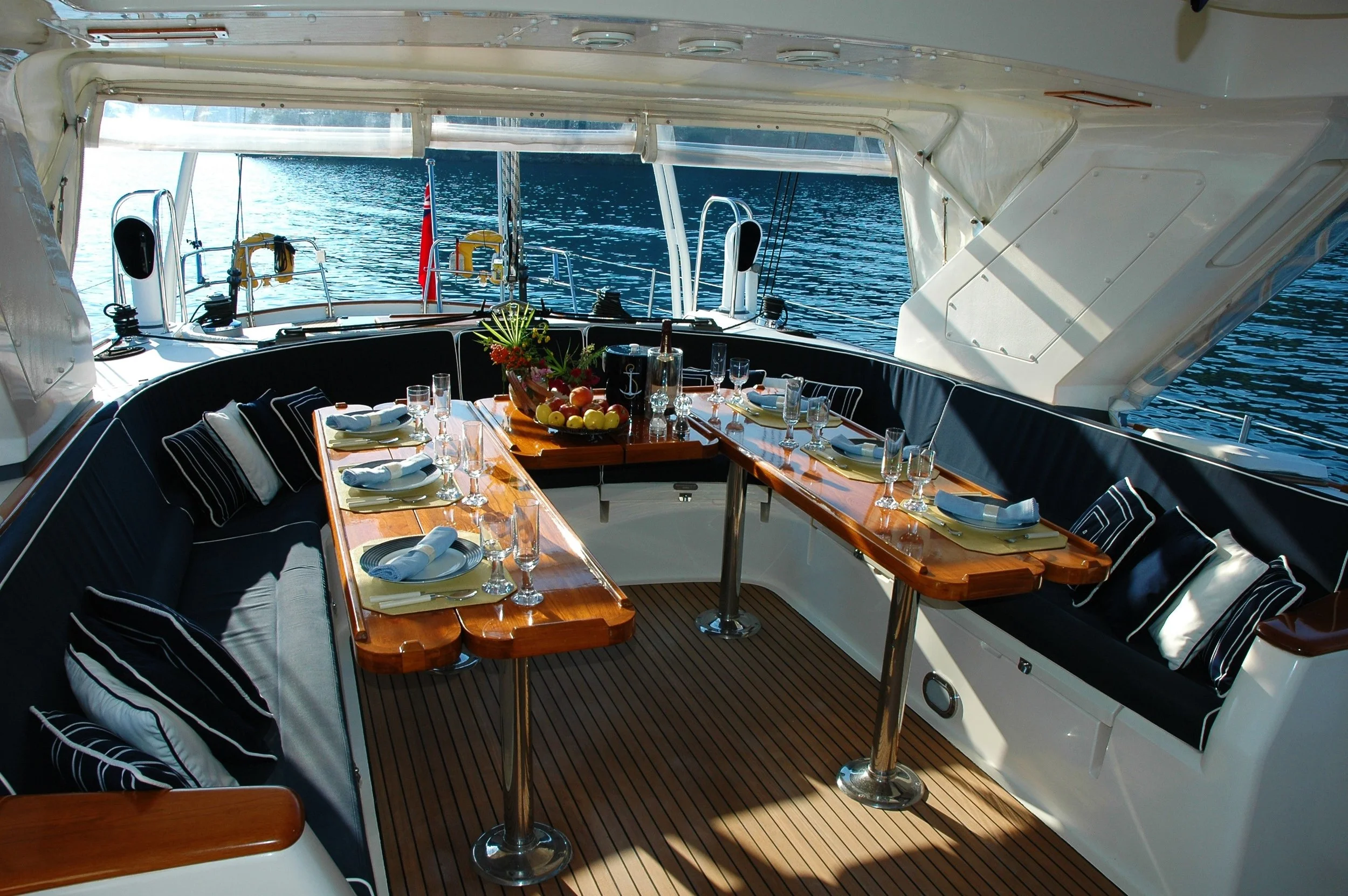 A luxurious yacht deck with a wooden dining table set with white plates, glasses, and napkins, surrounded by black cushioned seating with striped pillows, overlooking calm water.