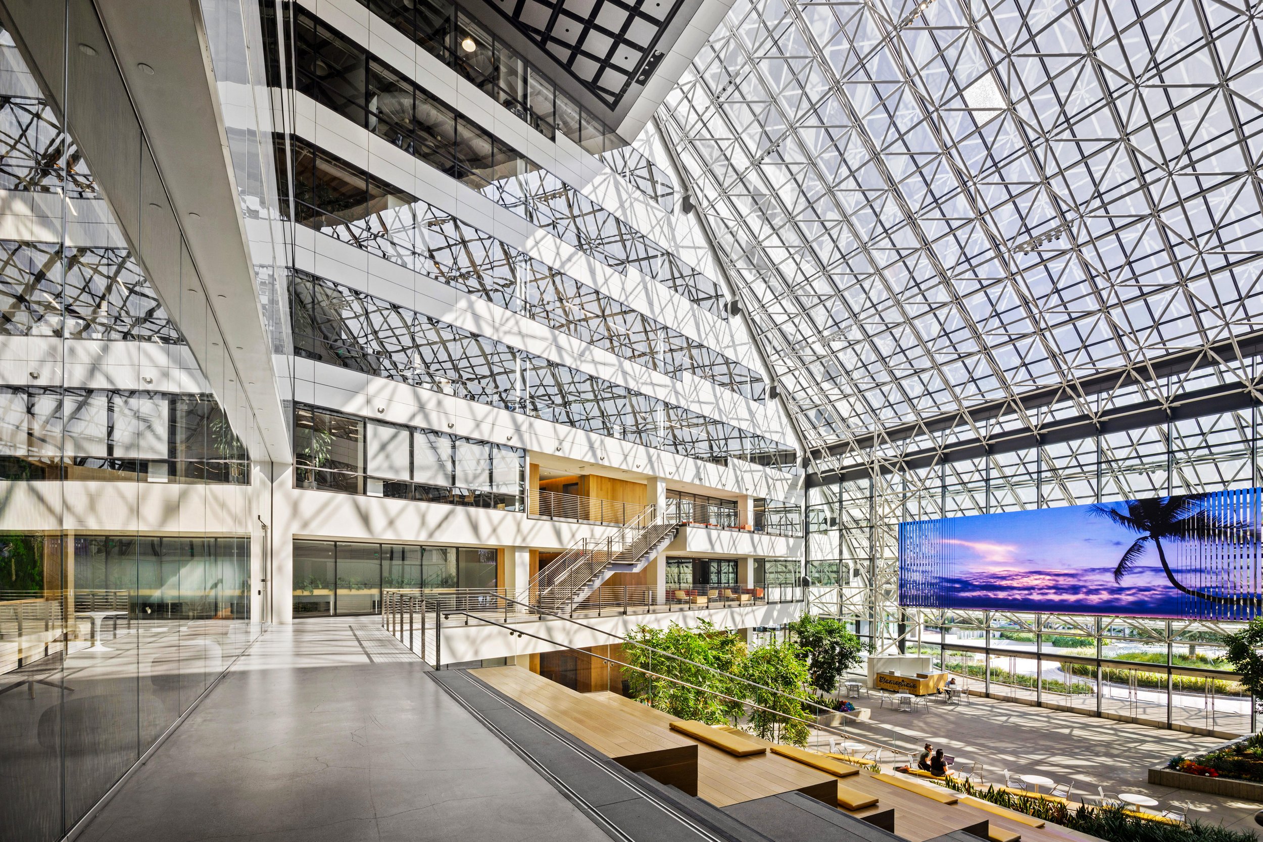 Interior of a modern building with a large glass and steel dome ceiling, multiple floors, glass walls, a large digital screen displaying a sunset scene, and seating areas with plants.