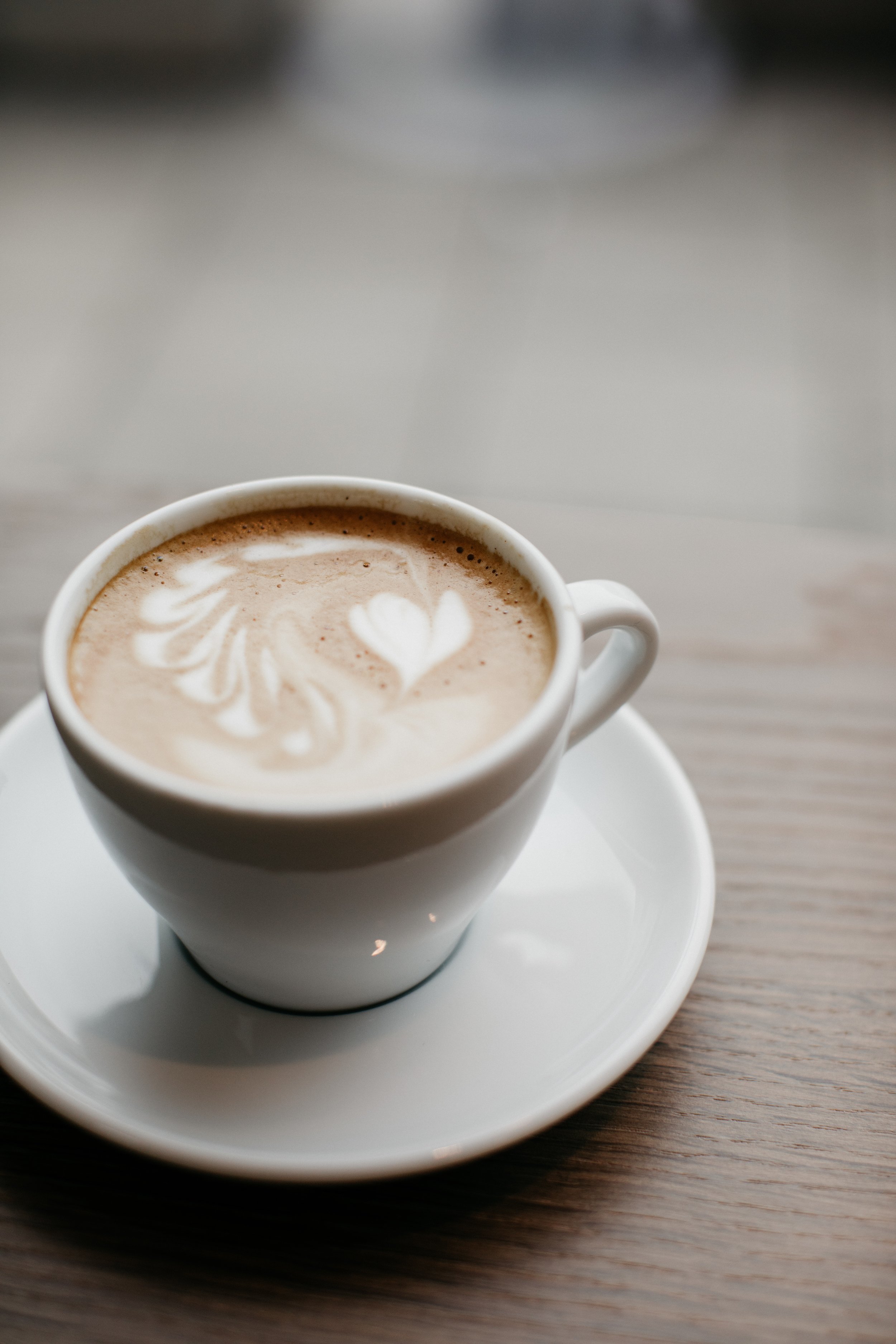 Kunstvoll zubereiteter Cappuccino mit Herz- und Blätter-Milchschaum-Design in einer weißen Tasse auf Holzschreibtisch.
