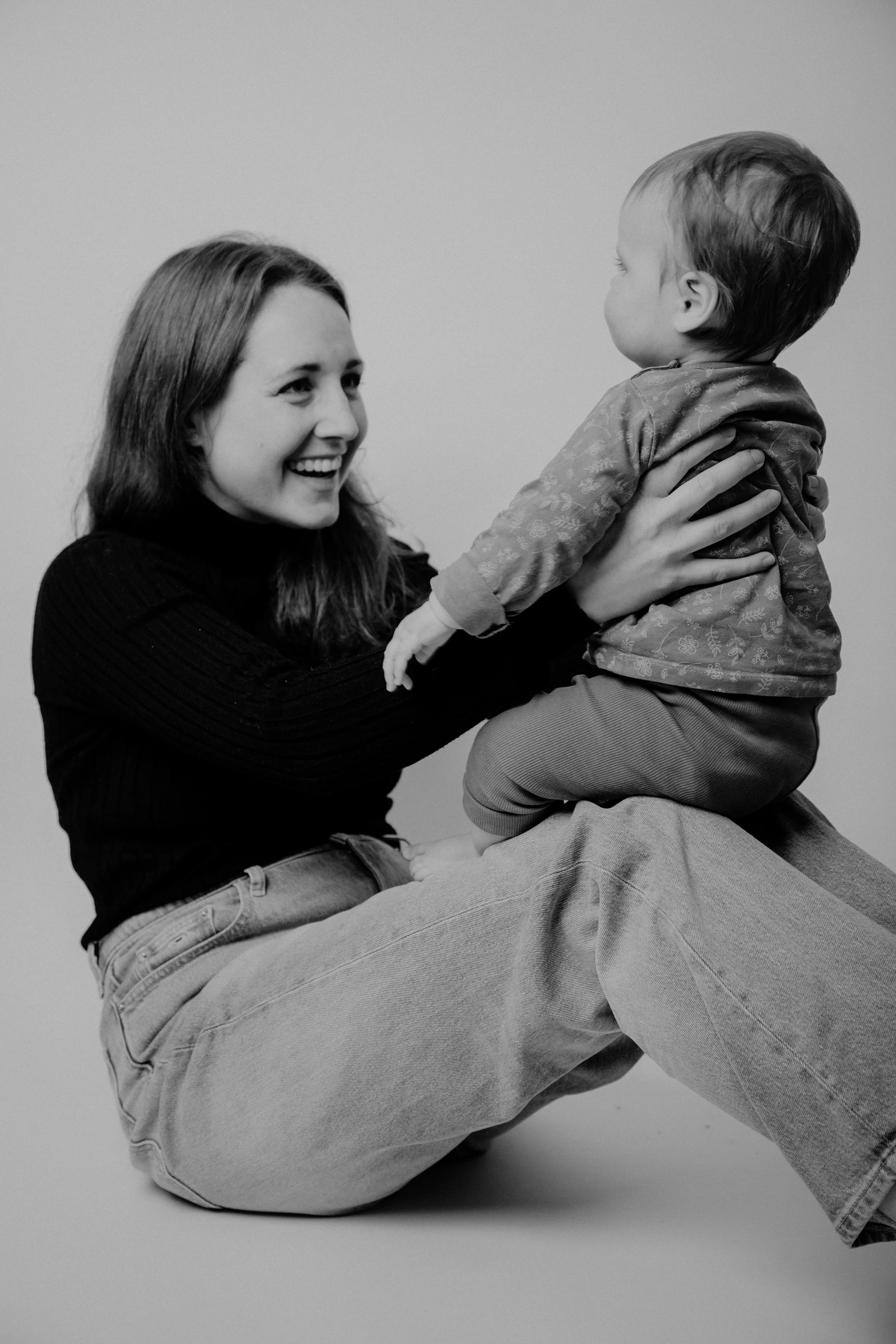 Eine junge Frau sitzt auf dem Boden, hält ein kleines Kind hoch und lächelt freundlich, beide sind schwarz-weiß fotografiert.