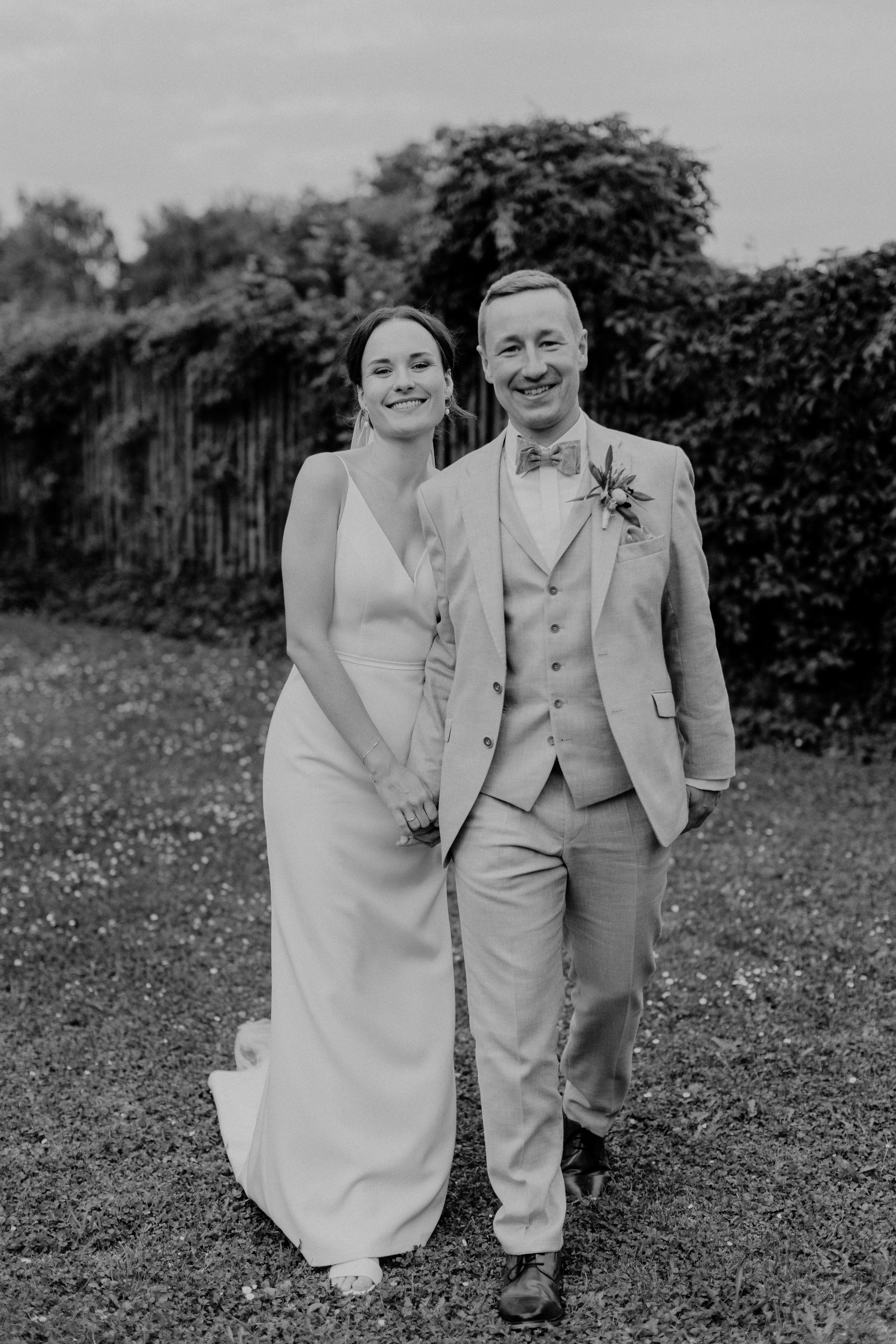 Ein lachendes Brautpaar in Hochzeitskleidung, die Hand in Hand draußen vor einer Hecke stehen.