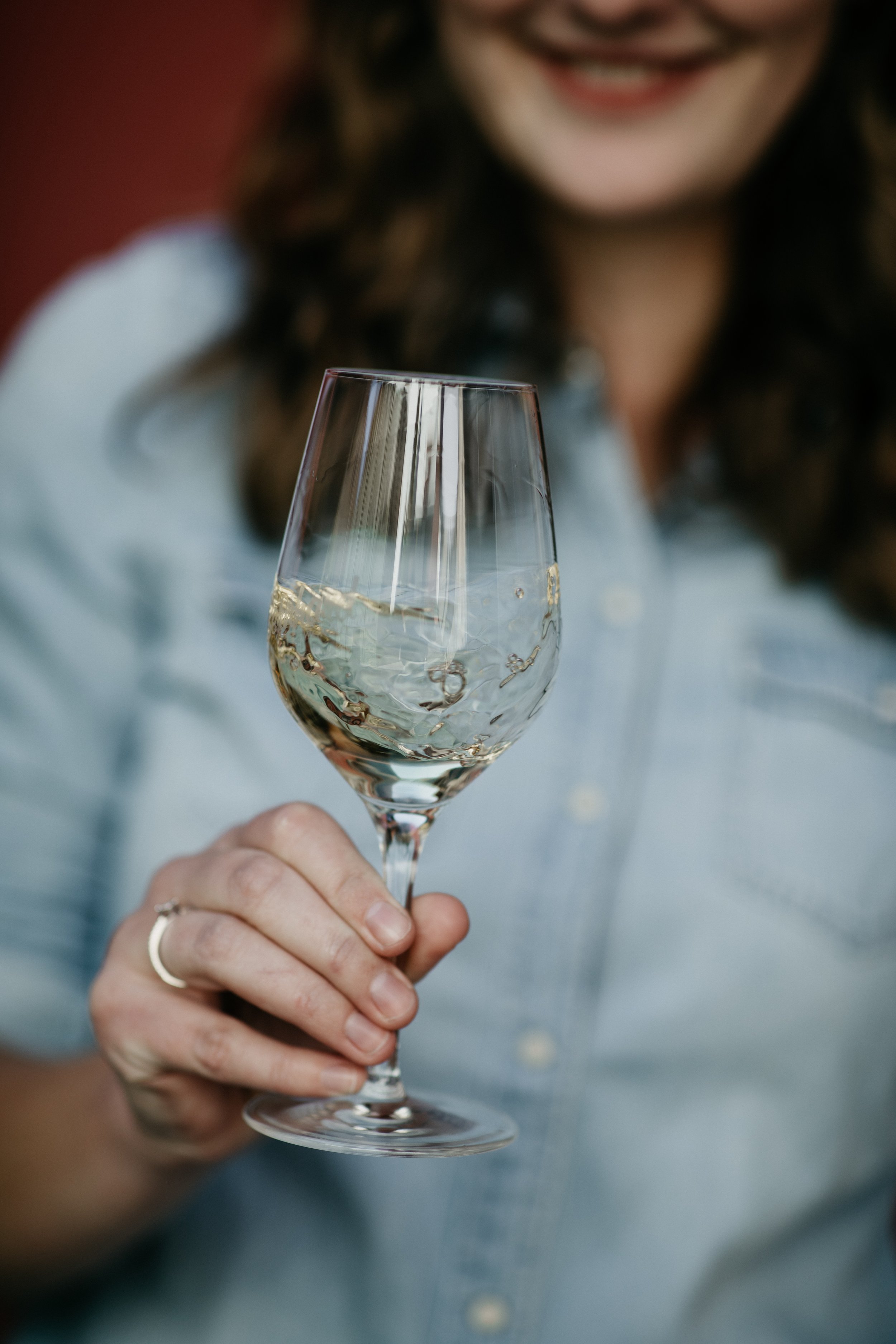 Frau hält ein Weinglas mit weißem Wein und lächelt im Hintergrund.