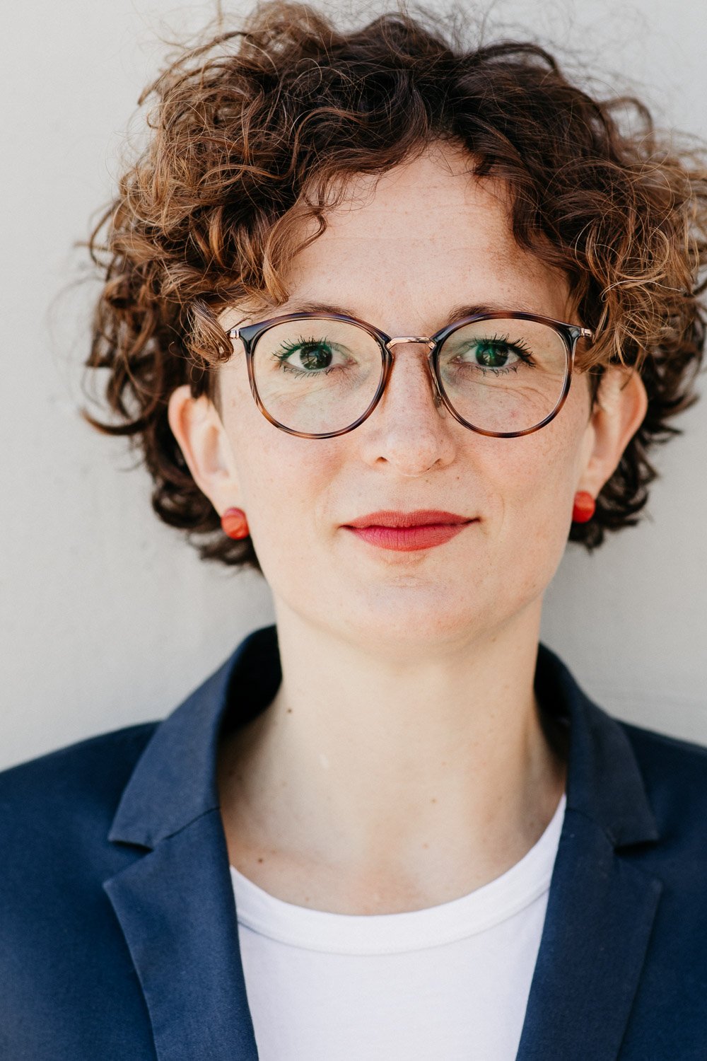 Nahaufnahme eines lachenden Frau mit kurzen, lockigen braunen Haaren, runden Brillen mit dunklem Rahmen, rotem Lippenstift, roter Bluse, dunkelblauer Jacke und roten Ohrsteckern vor hellem Hintergrund.