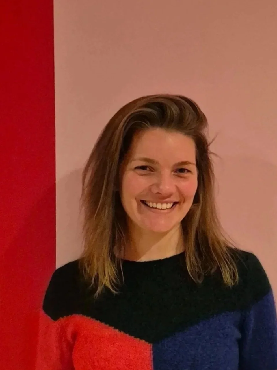 Een glimlachende vrouw met bruin haar, gekleed in een zwart, blauw en rood geblokt trui, tienertoussig voor een roze en rode muur.