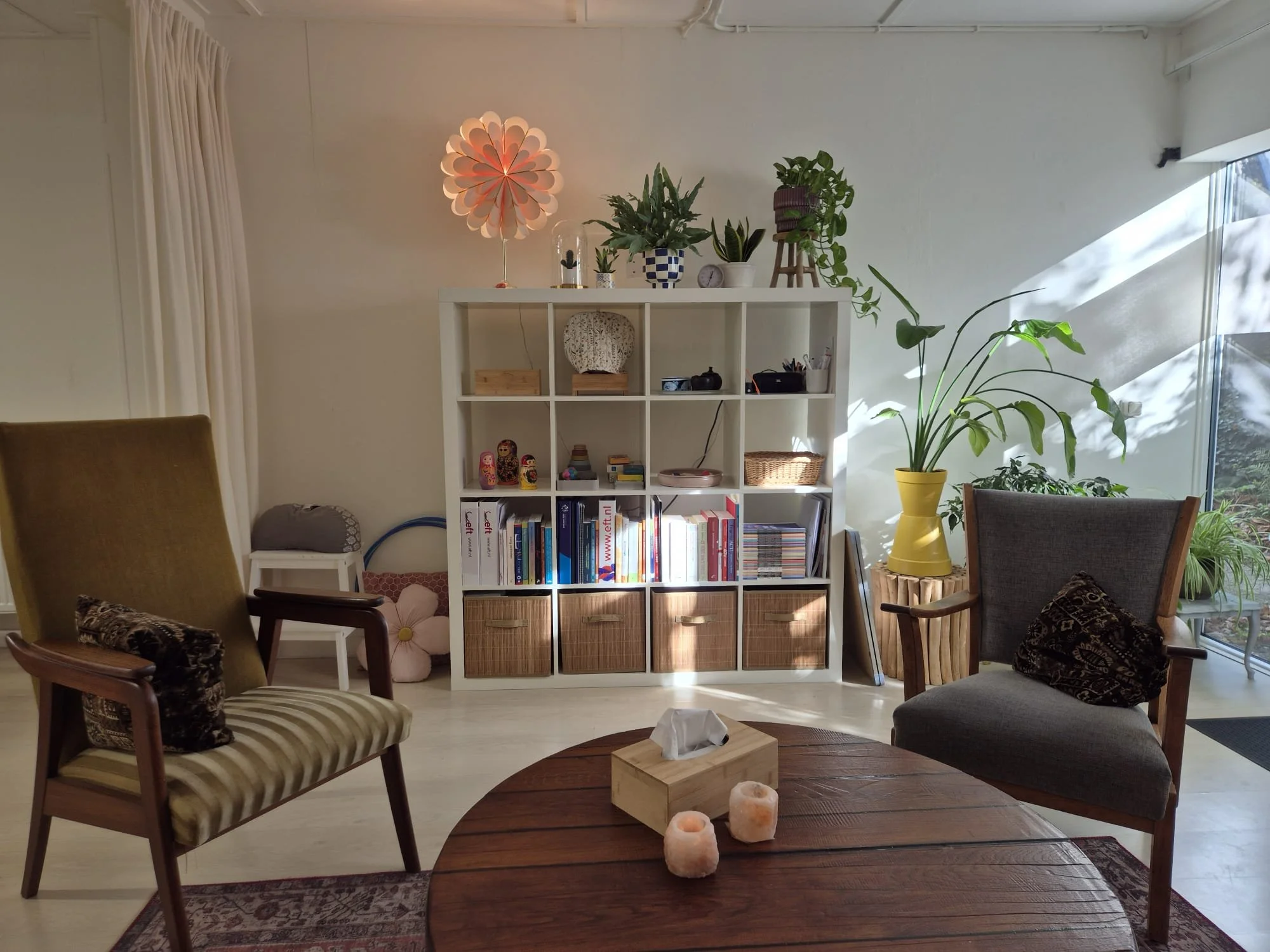 Woonkamer met twee fauteuils, een ronde houten tafel met een doos tissues en drie kaarsen, een witte boekenplank gevuld met boeken en decoratieve items, en planten op en naast de plank, met grote ramen aan de rechterkant waar zonlicht binnenkomt.