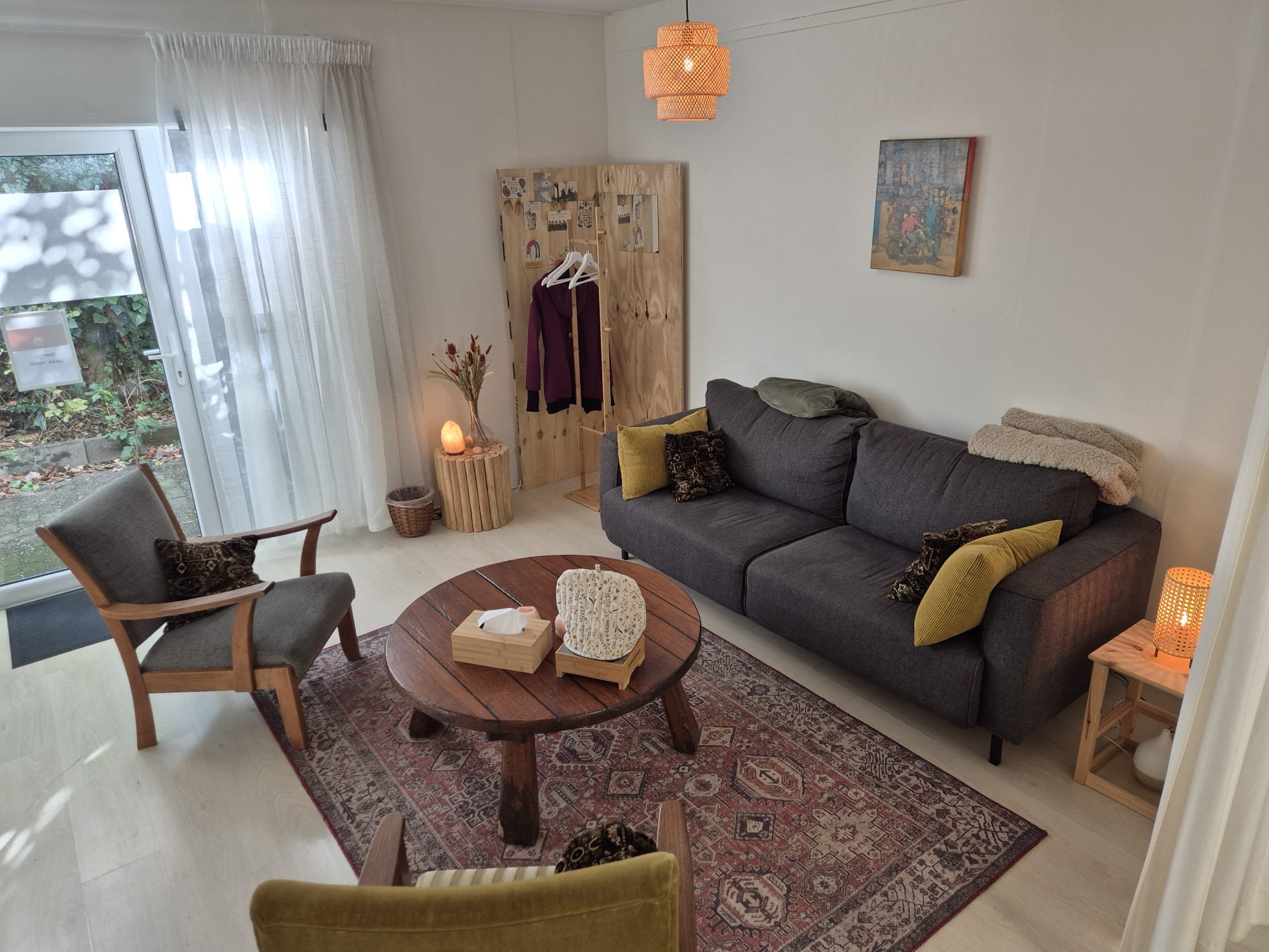 Een woonkamer met een donkergrijze bank, een houten salontafel, een armstoel, een vloerkleed, een grote raam met witte gordijnen, een lamp, een schilderij aan de muur en een kledingrek.