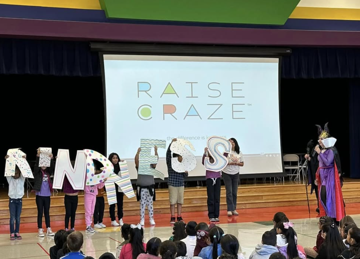 Children on stage holding decorated letters spelling 'RND' with a woman dressed as a witch or fairy standing to the right, speaking into a microphone, in front of an audience in a gymnasium. There is a large screen behind them displaying the 'Raise Craze' logo.