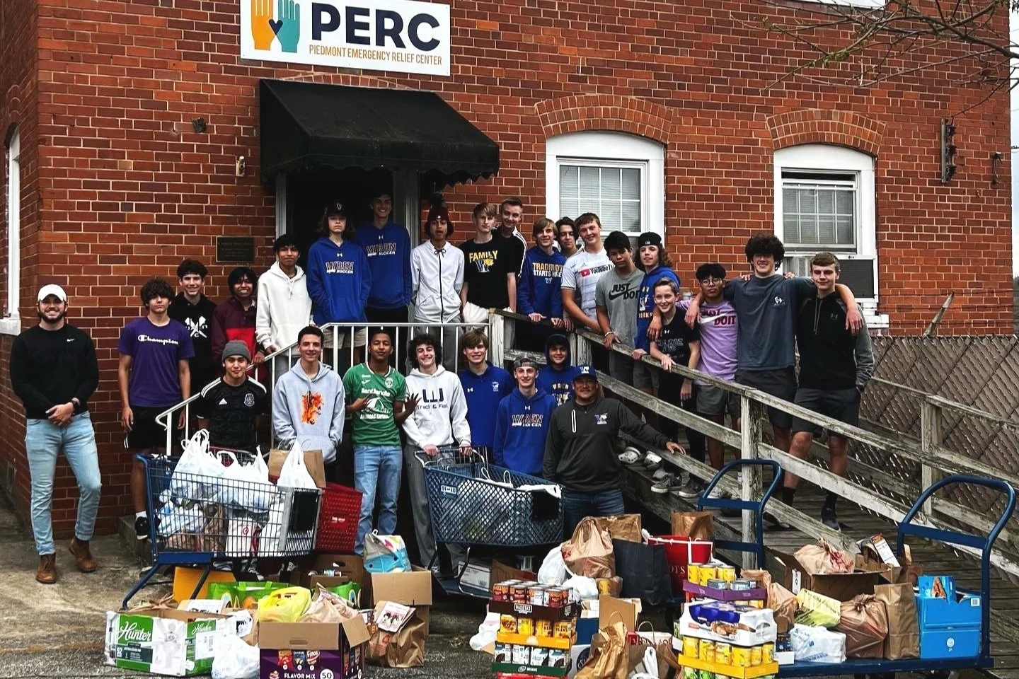 Group of people standing on the front steps of a brick building with a sign that reads 'PERC Piedmont Emergency Relief Center.' There are shopping carts filled with groceries in front of them.