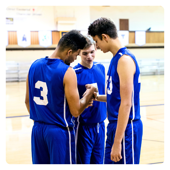 Three young male basketball players in blue jerseys standing in a gymnasium, huddling and looking at a phone held by one of them.