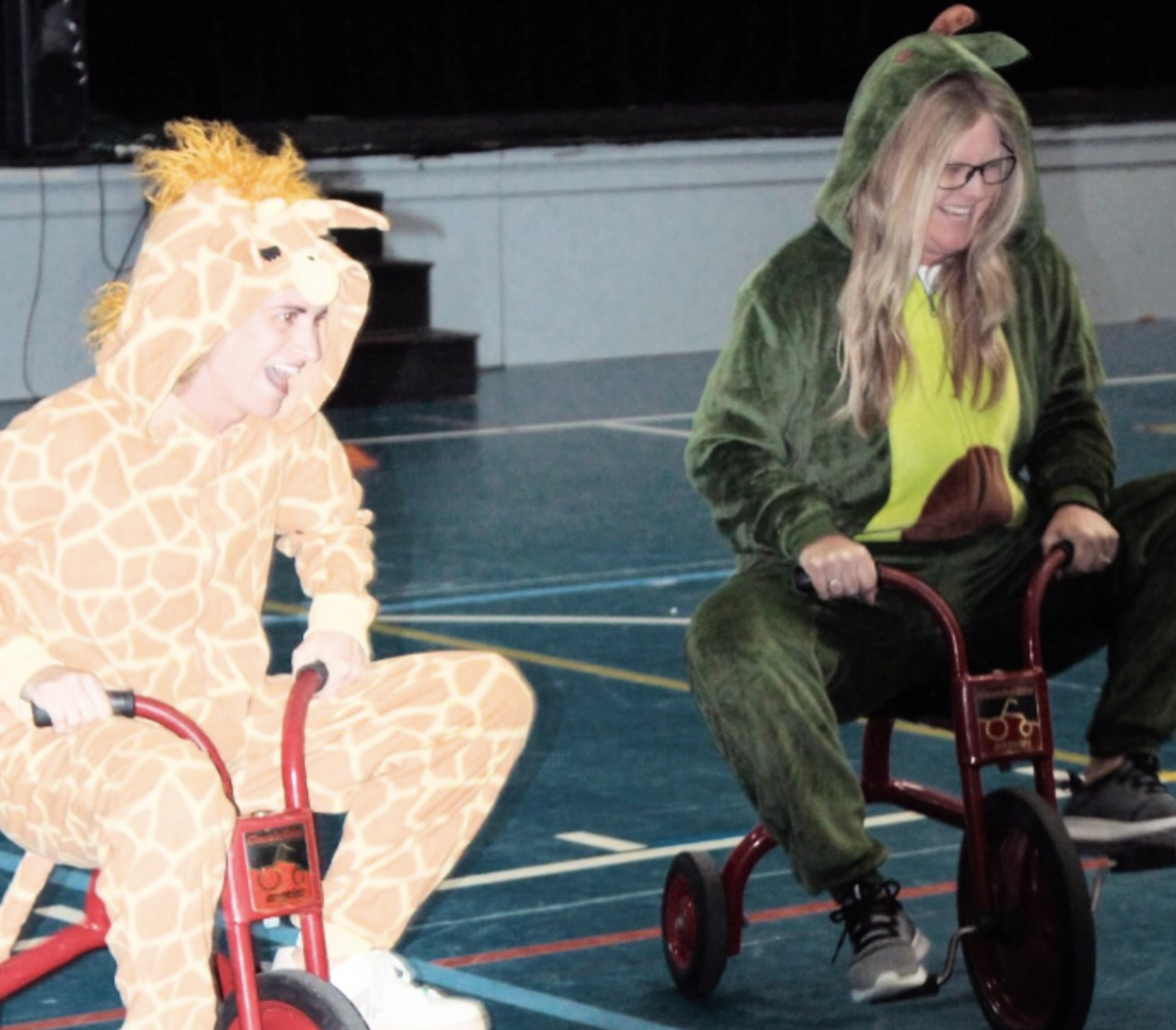 Two women dressed in animal costumes, a giraffe and a crocodile, riding tricycles on a gymnasium floor, smiling and having fun.