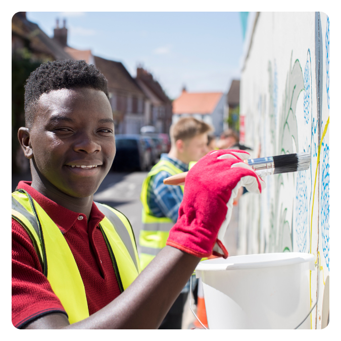 A young boy with dark skin, wearing a red shirt and yellow safety vest, is painting a mural on a wall with a large paintbrush. He is smiling at the camera, and other children are painting in the background on a sunny day.