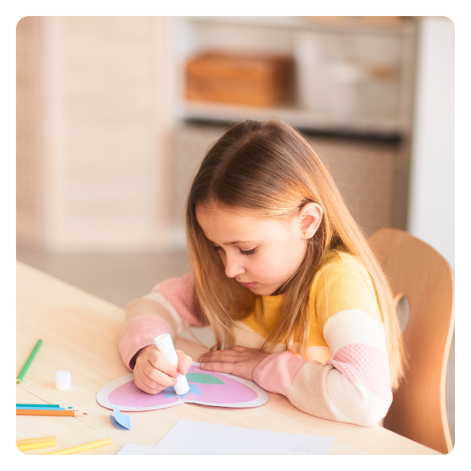 A young girl with long light brown hair is sitting at a table, coloring a paper with a white glue stick. She is wearing a yellow and pink long-sleeved shirt and appears focused on her craft project.