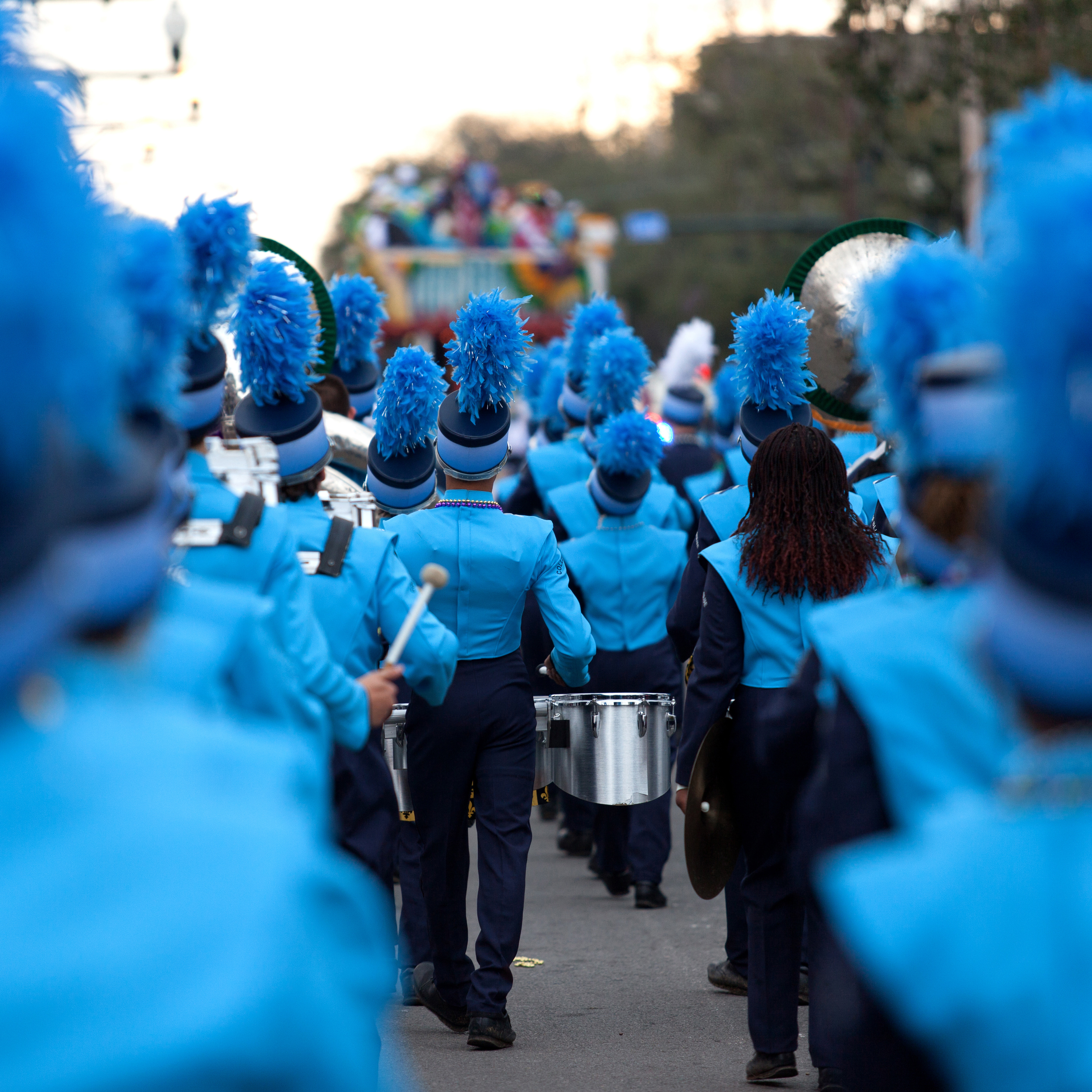 A marching band dressed in blue costumes with blue feathered hats, playing instruments as they march down a street during a parade at dusk.