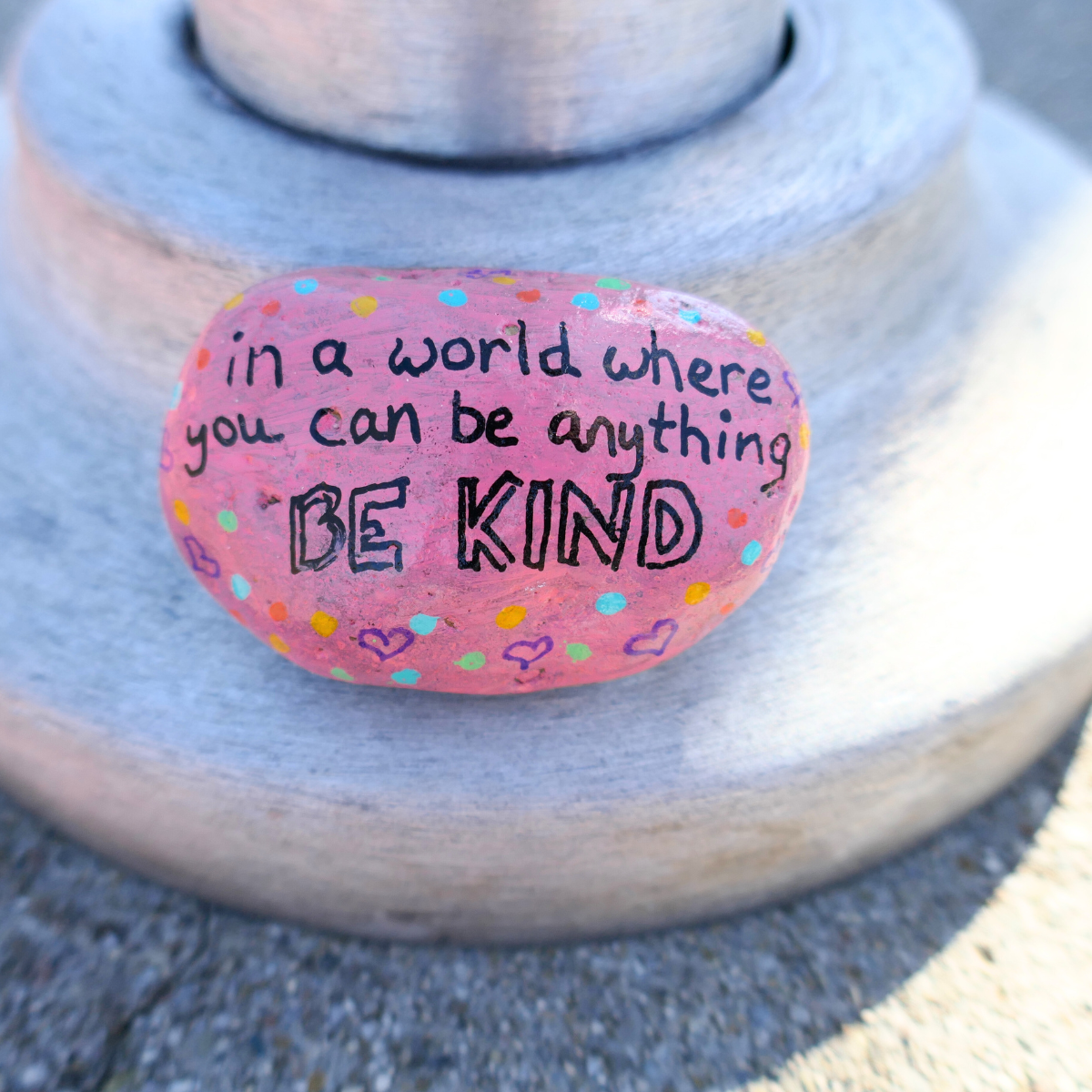 Pink painted rock with colorful dots and the message 'in a world where you can be anything, BE KIND' written on it, placed on a gray surface.