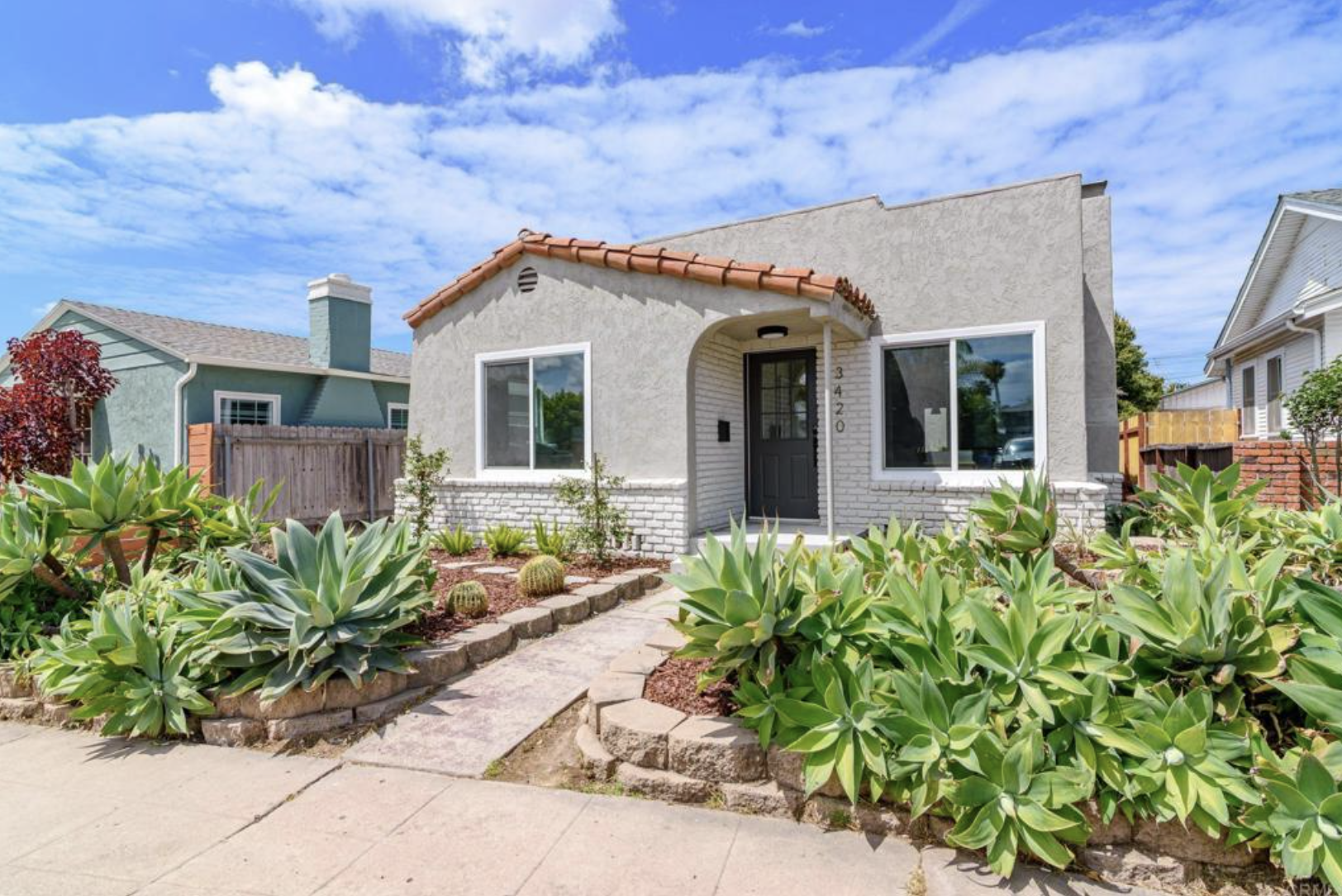 Single-story house with a landscaped front yard featuring large green succulents and small cacti, a concrete pathway leading to the black front door, and neighboring houses on a sunny day with a partly cloudy sky.
