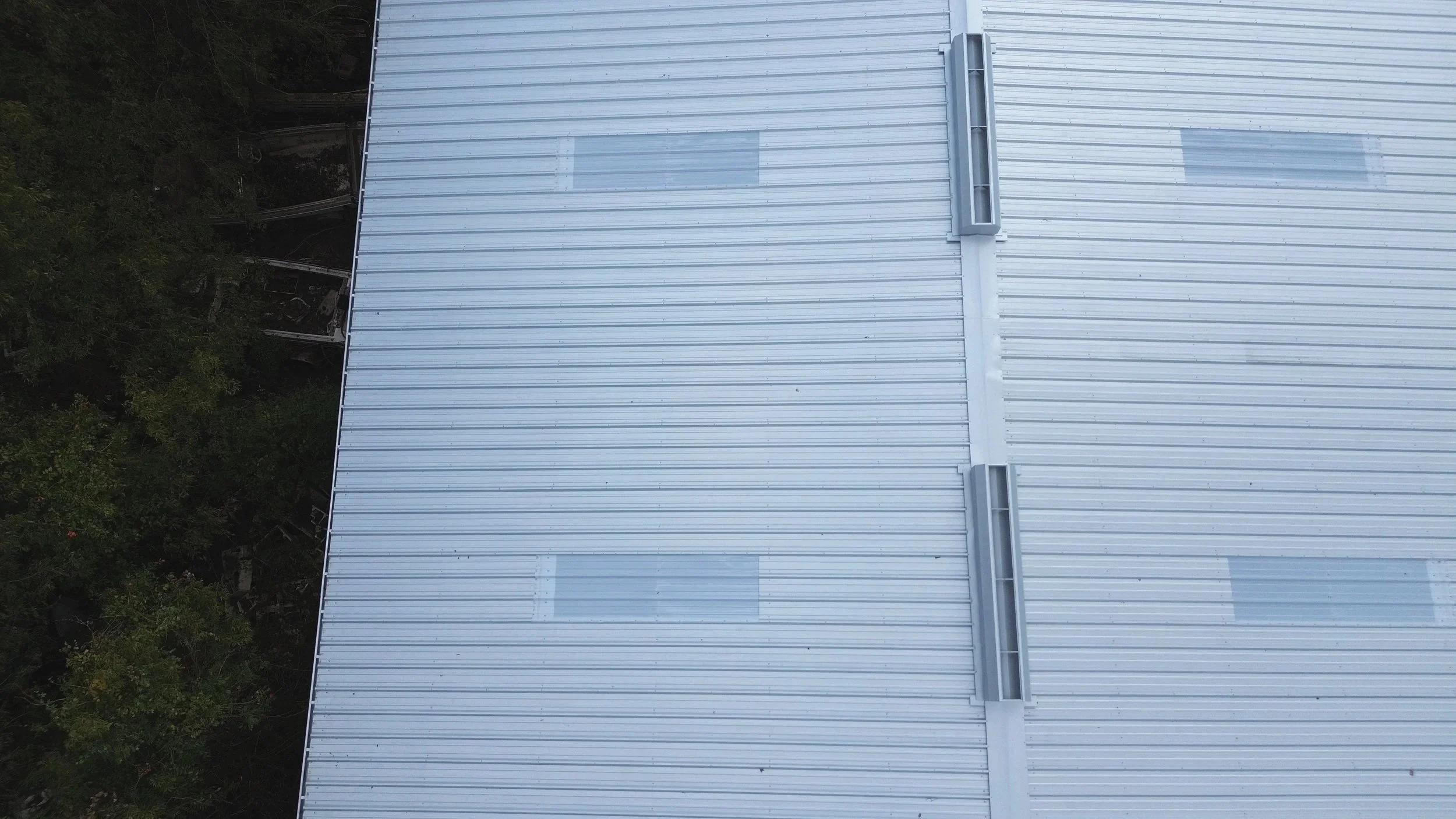Overhead view of a metal rooftop with vents and skylights, surrounded by trees on the left side.