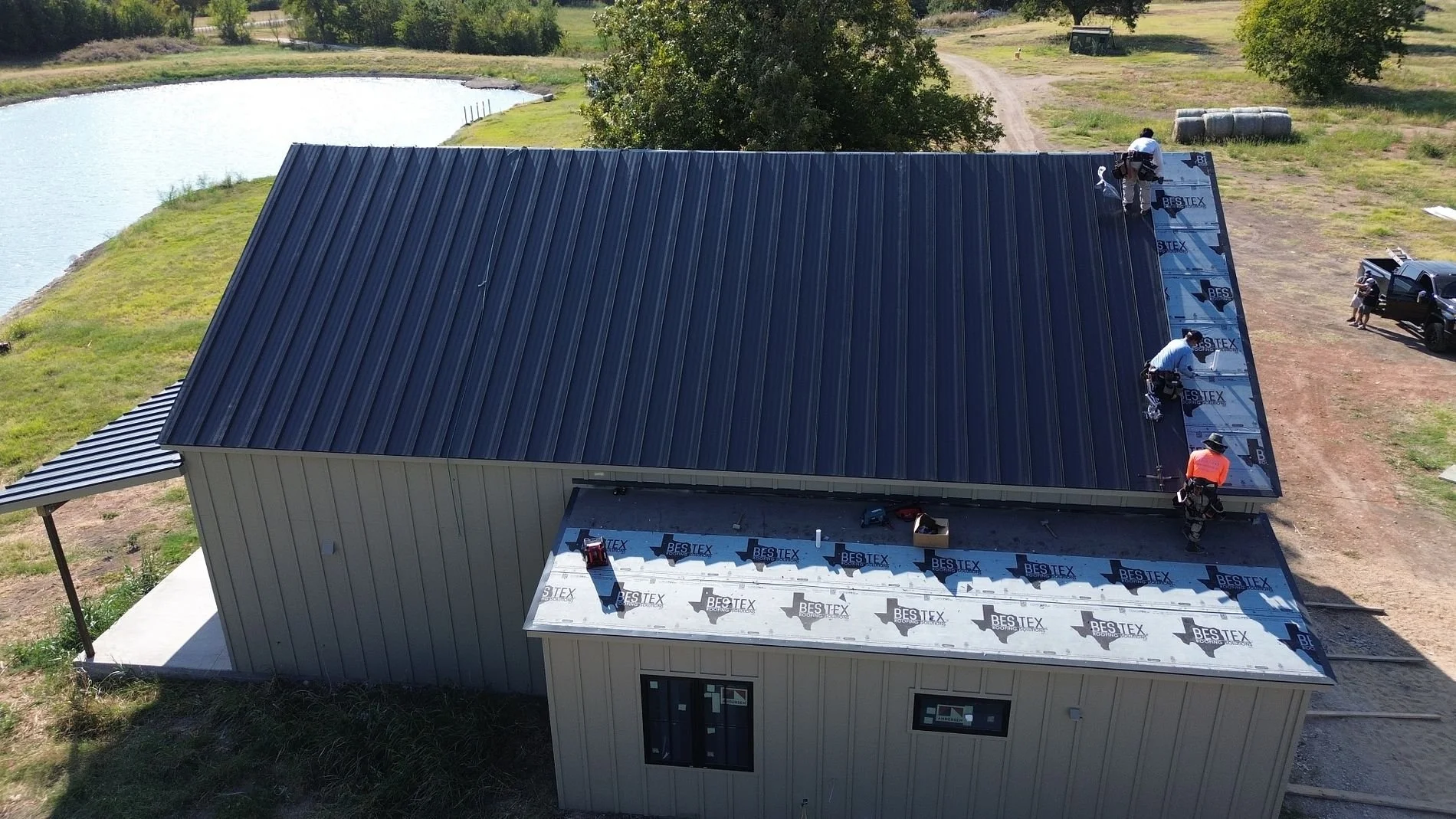 Two workers installing roof sheathing and waterproof membrane on a sloped roof during construction, with trees and a road visible in the background.