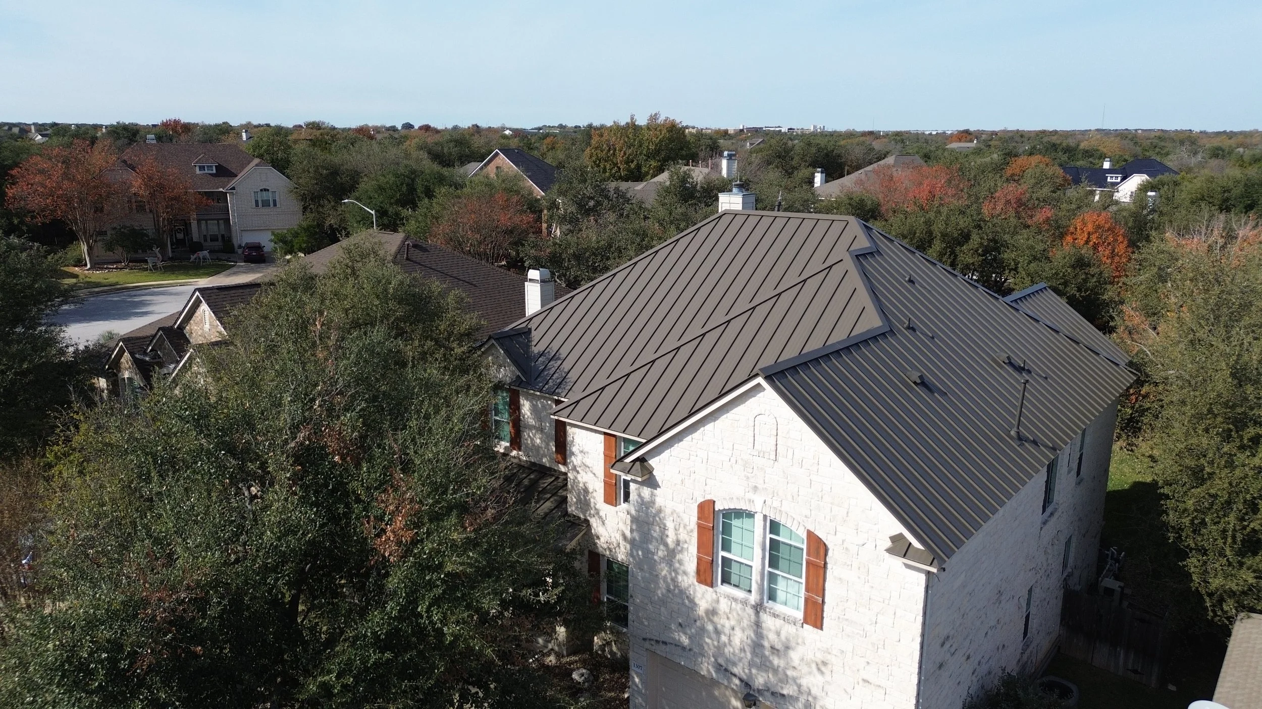 An aerial view of a house with a black metal roof, surrounded by trees and a grassy yard, with a patio and garden area visible.