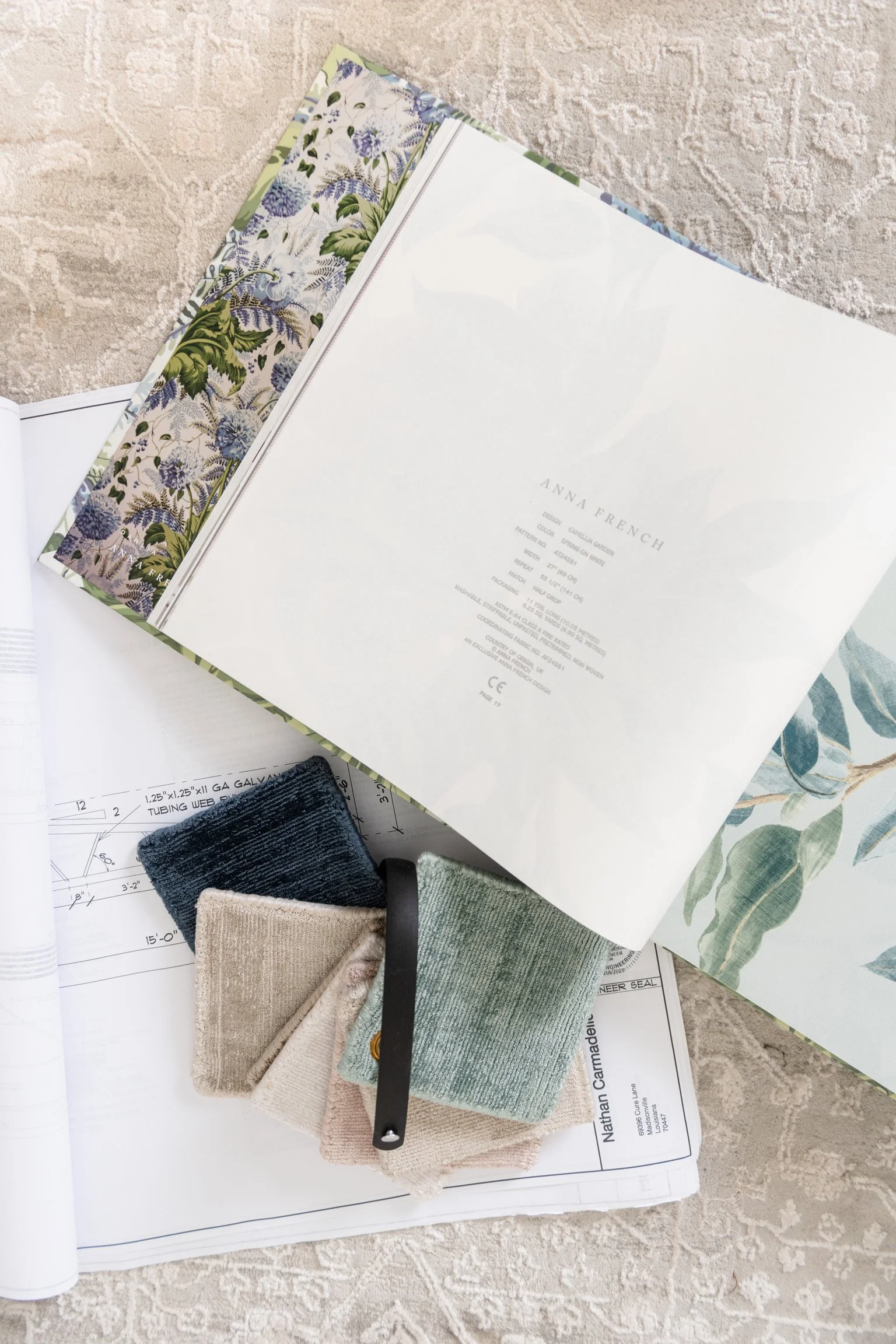Interior design samples including a large open book with leaf and floral patterns, paint swatches in beige, green, and dark blue, and architectural blueprints on a beige carpet.