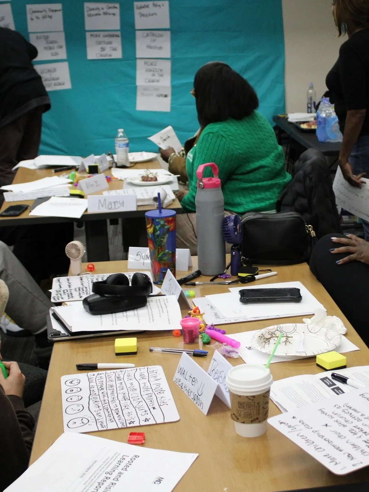 A group of people seated at a table engaged in a workshop or meeting, with papers, notebooks, snacks, and personal items scattered across the table. There is a blue bulletin board or poster in the background with notes pinned to it.