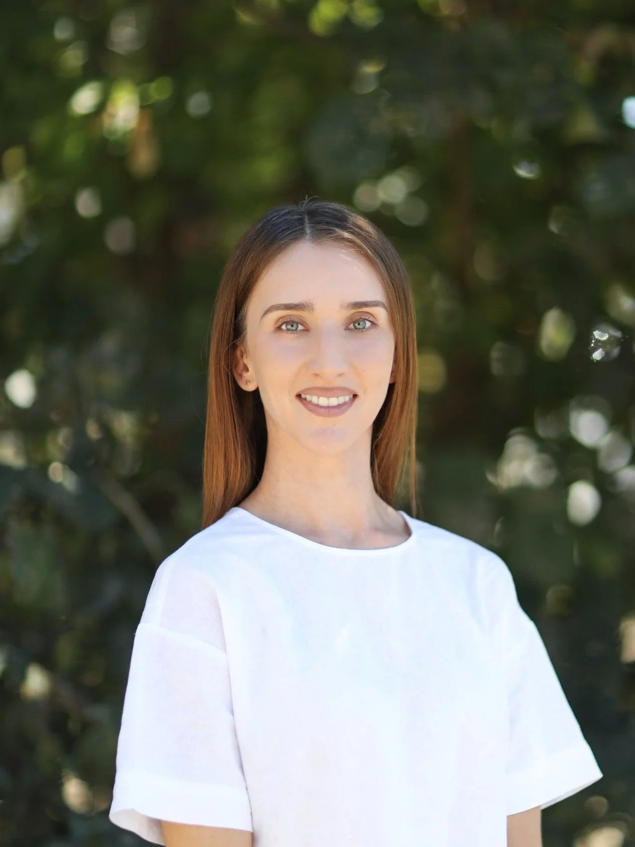 A woman with long light brown hair, blue eyes, and fair skin, wearing a white shirt, standing outdoors in front of a background of blurred green foliage.