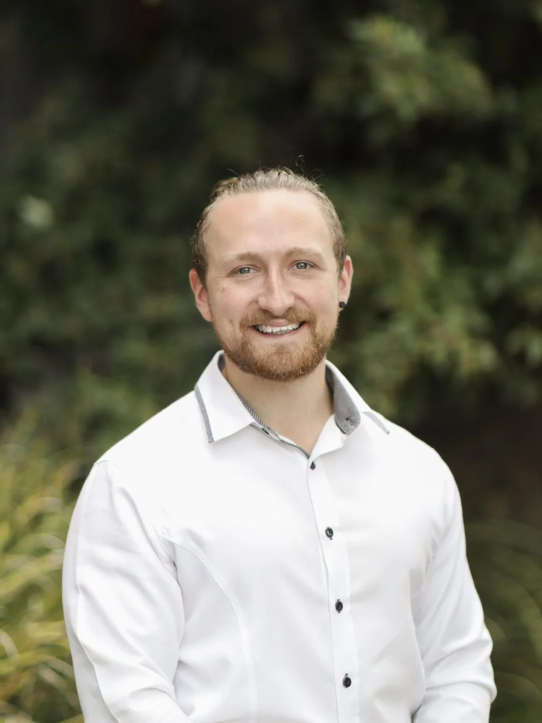 Portrait of a smiling man with blond hair and a beard, wearing a white button-up shirt, standing outdoors with blurred green foliage in the background.