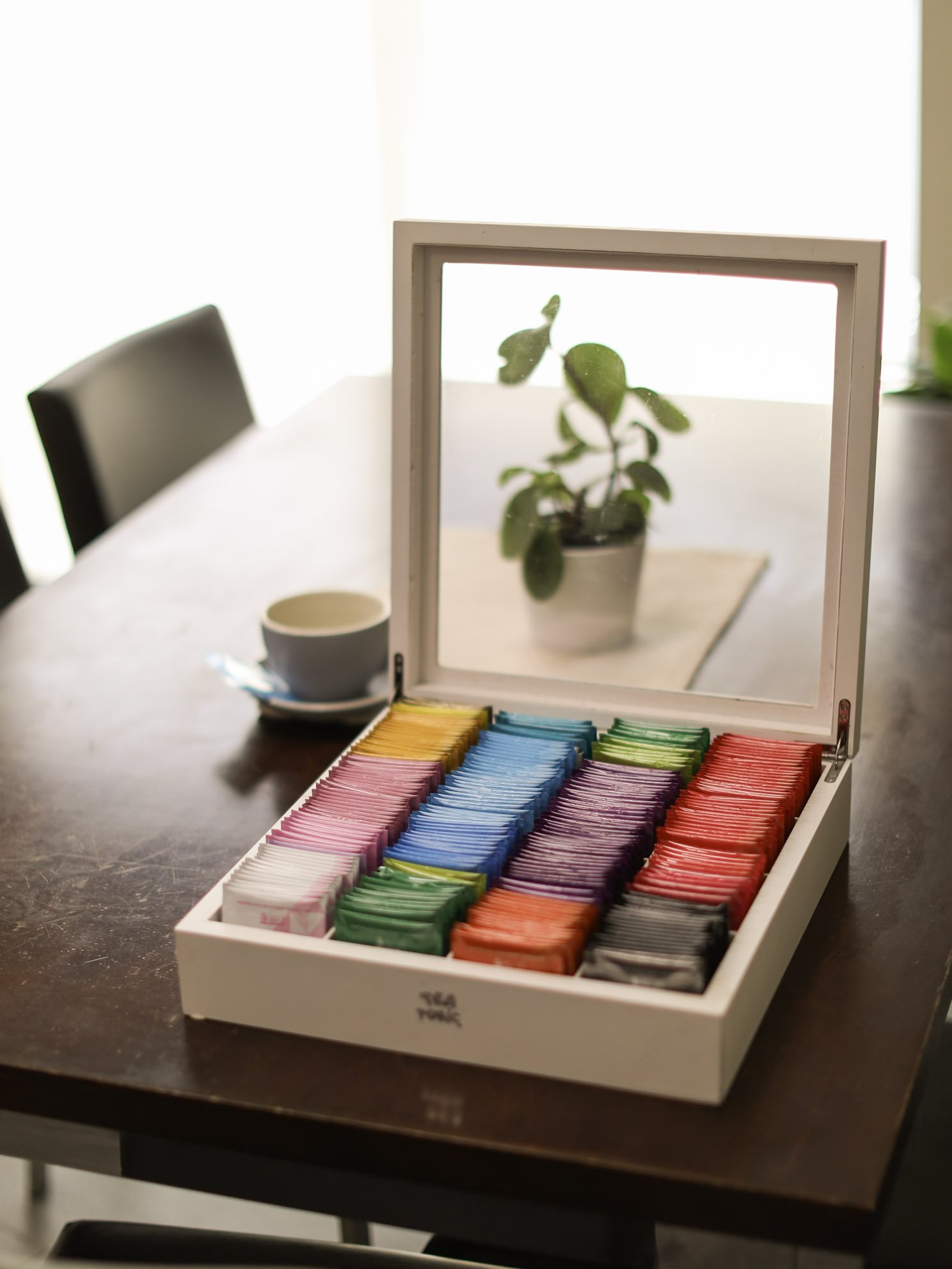 A white box with colored sugar packets is open on a dark wooden table, with a mirror on the inside of the lid reflecting a potted plant in the background.