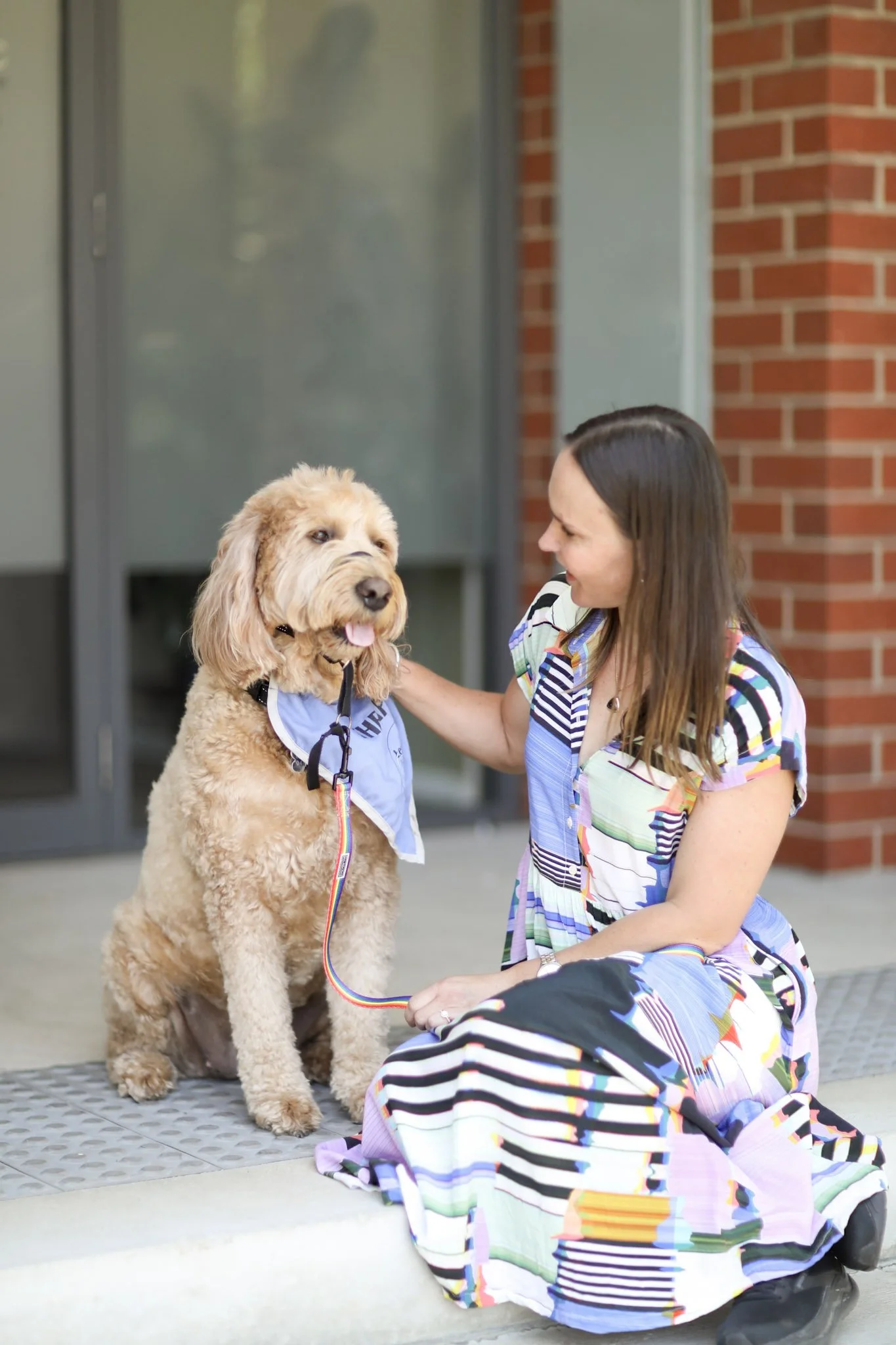 A woman kneeling and petting a large, cream-colored dog sitting on a porch in front of a brick wall and glass door.