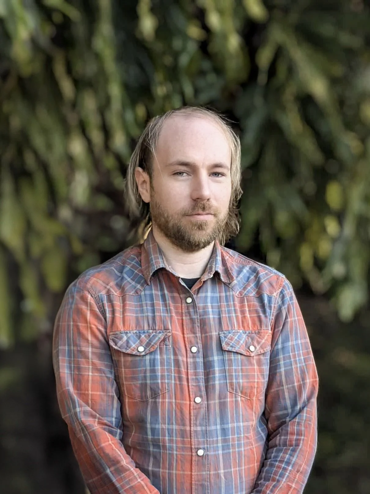 A man with long hair and a beard standing outdoors in front of a background of green moss-covered rocks, wearing a plaid shirt.