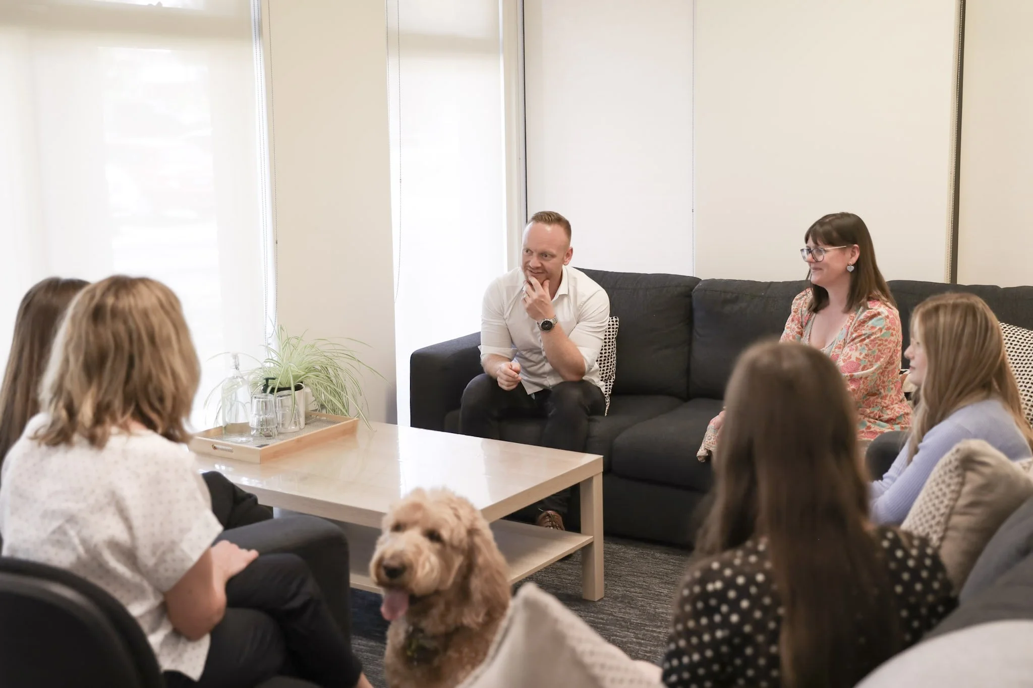 A group of people in a casual meeting room listening to a man speaking, with a dog in the foreground.