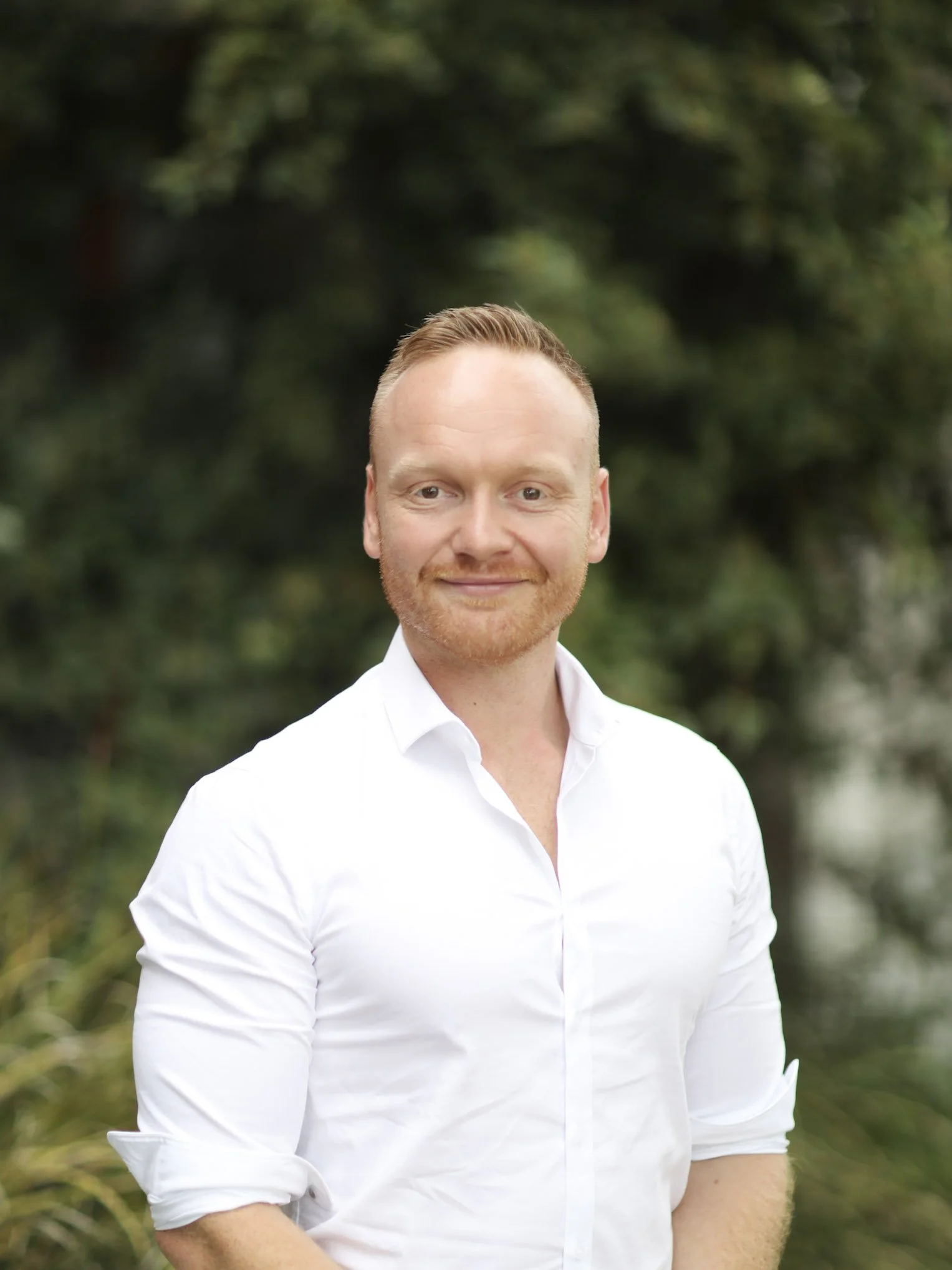 A man with red hair and beard, wearing a white shirt, smiling outdoors with a blurred green background.