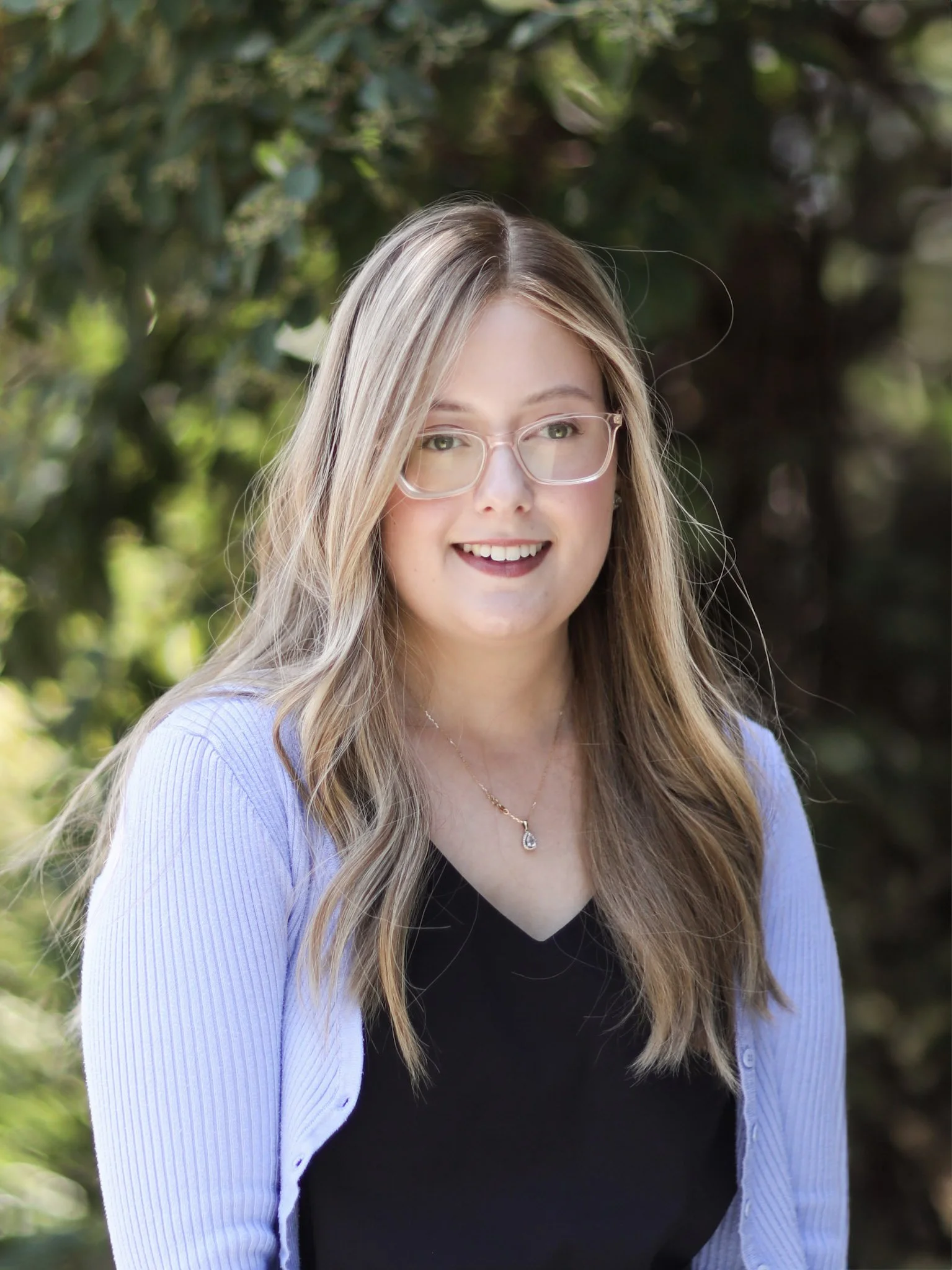 A young woman with blonde hair, glasses, and a necklace, smiling outdoors with greenery in background.