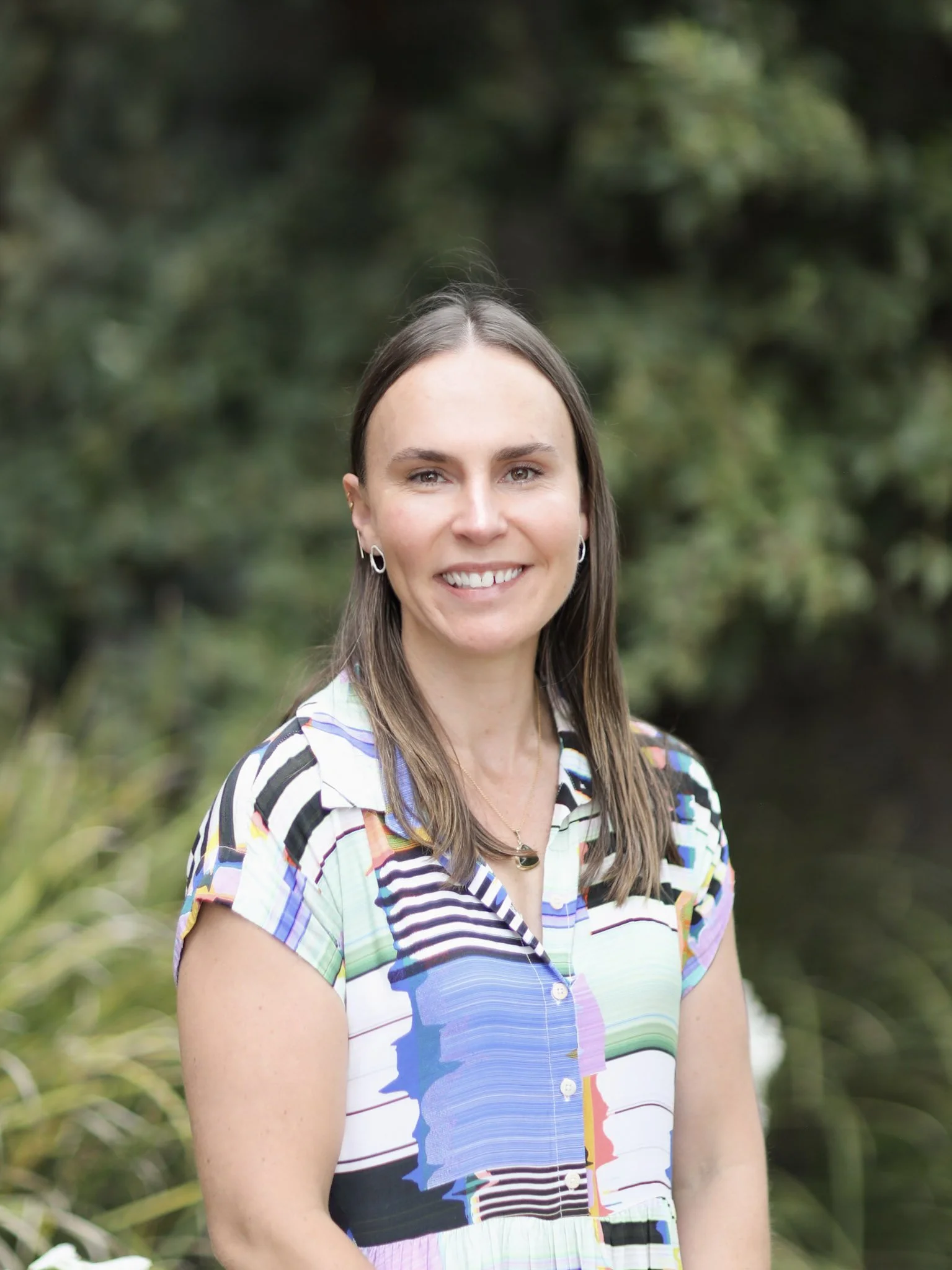 A woman with long brown hair smiling outdoors, wearing a colorful patterned blouse and earrings, with greenery in the background.
