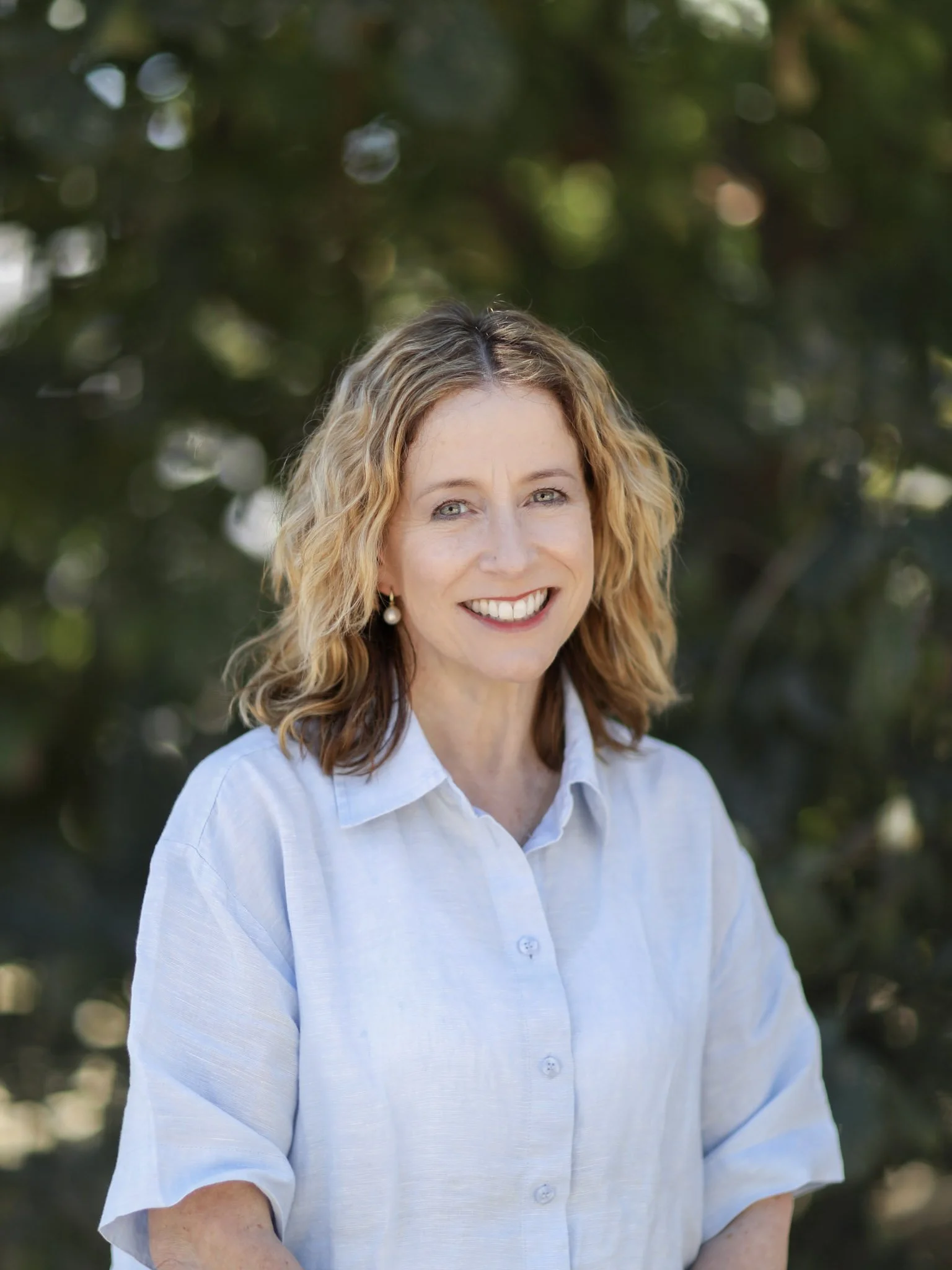 Smiling woman with shoulder-length curly blonde hair, wearing a white button-up shirt, outdoors with blurred greenery background.