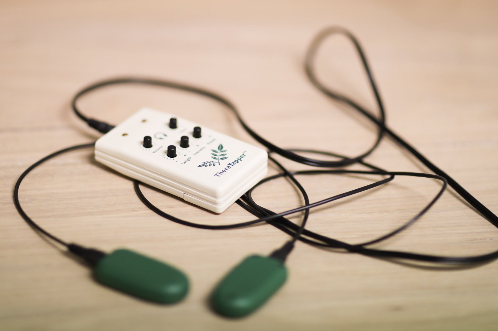 A white electronic device with black knobs labeled 'TheraTapper' connected to green massage applicators via black wires, placed on a light wooden surface.