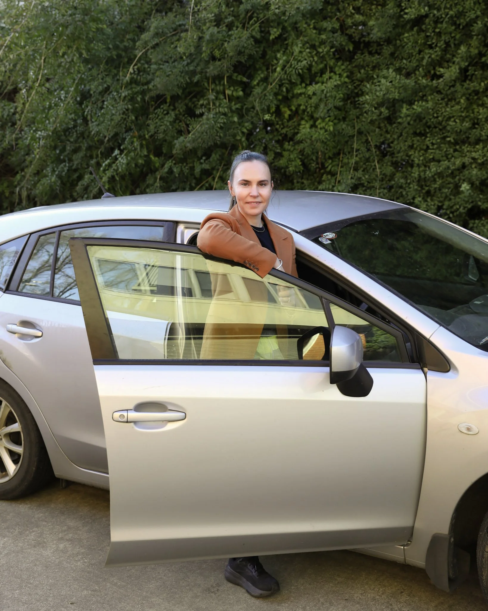 A woman with dark hair standing next to a silver car, resting her arms on the open driver's side door, with green trees in the background.