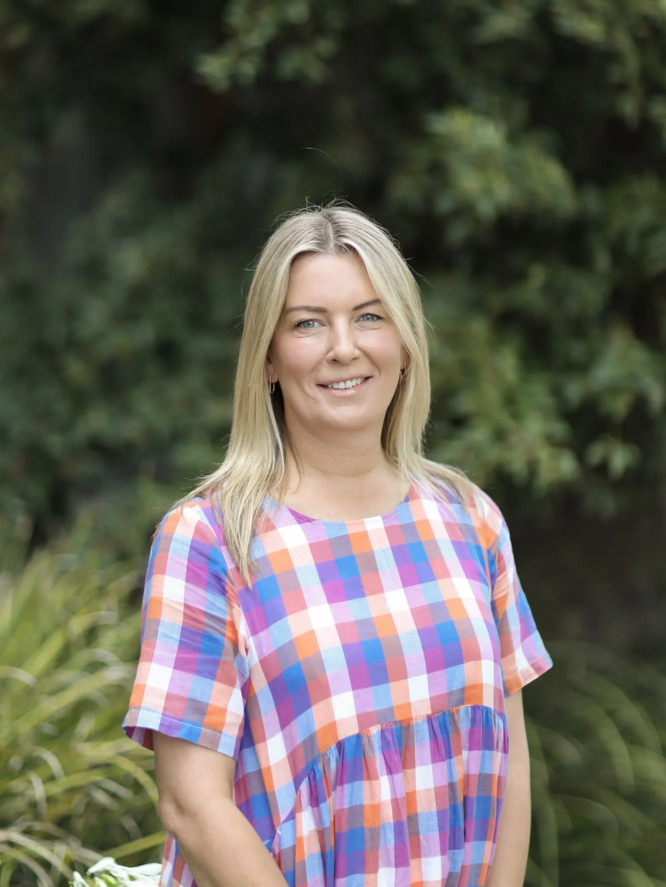 A woman with blonde hair wearing a multicolored plaid dress stands outdoors in front of green foliage, smiling at the camera.