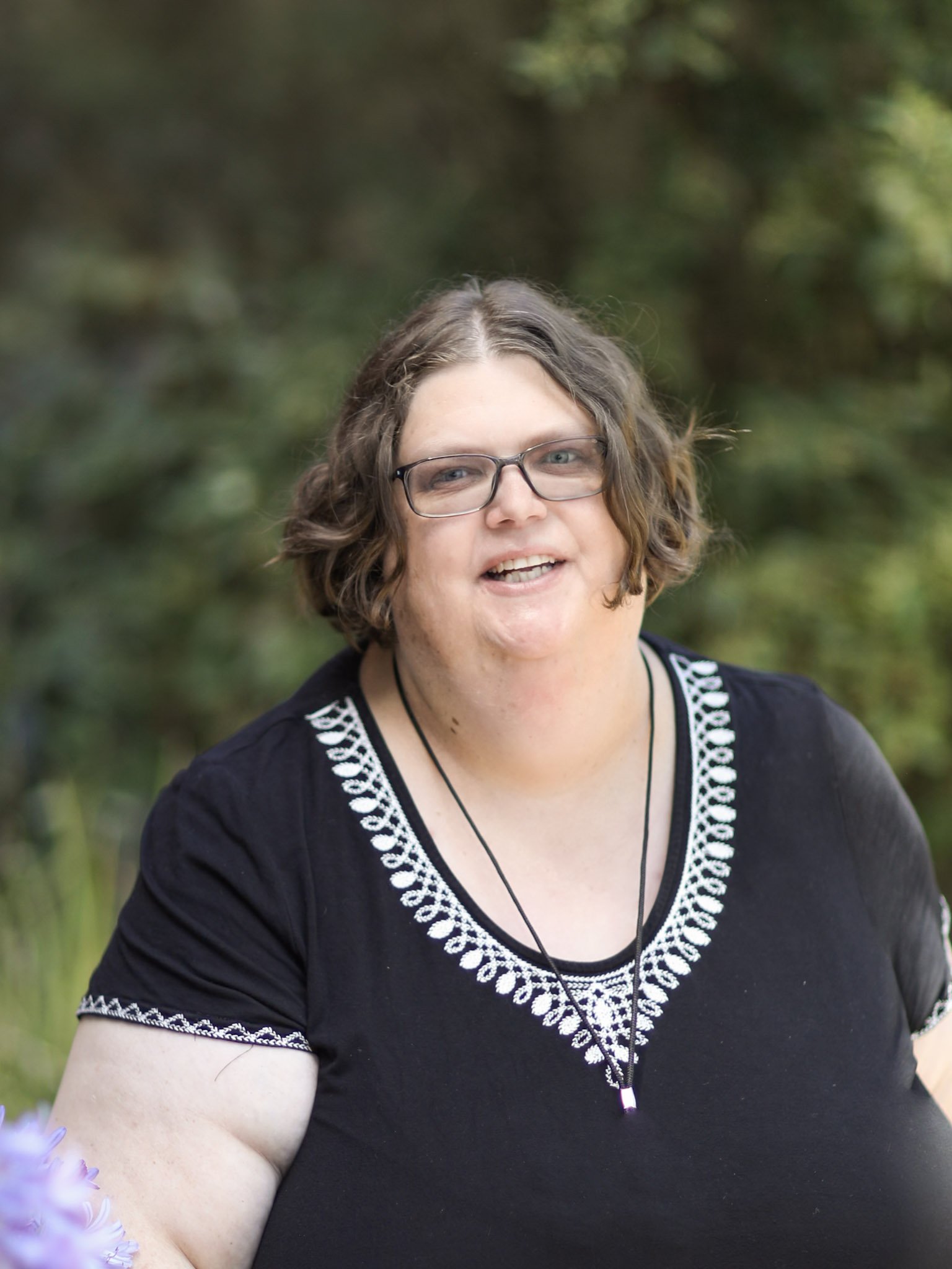 A woman with glasses, short curly brown hair, wearing a black top with white embroidery, and a necklace, outdoors with green foliage in the background.