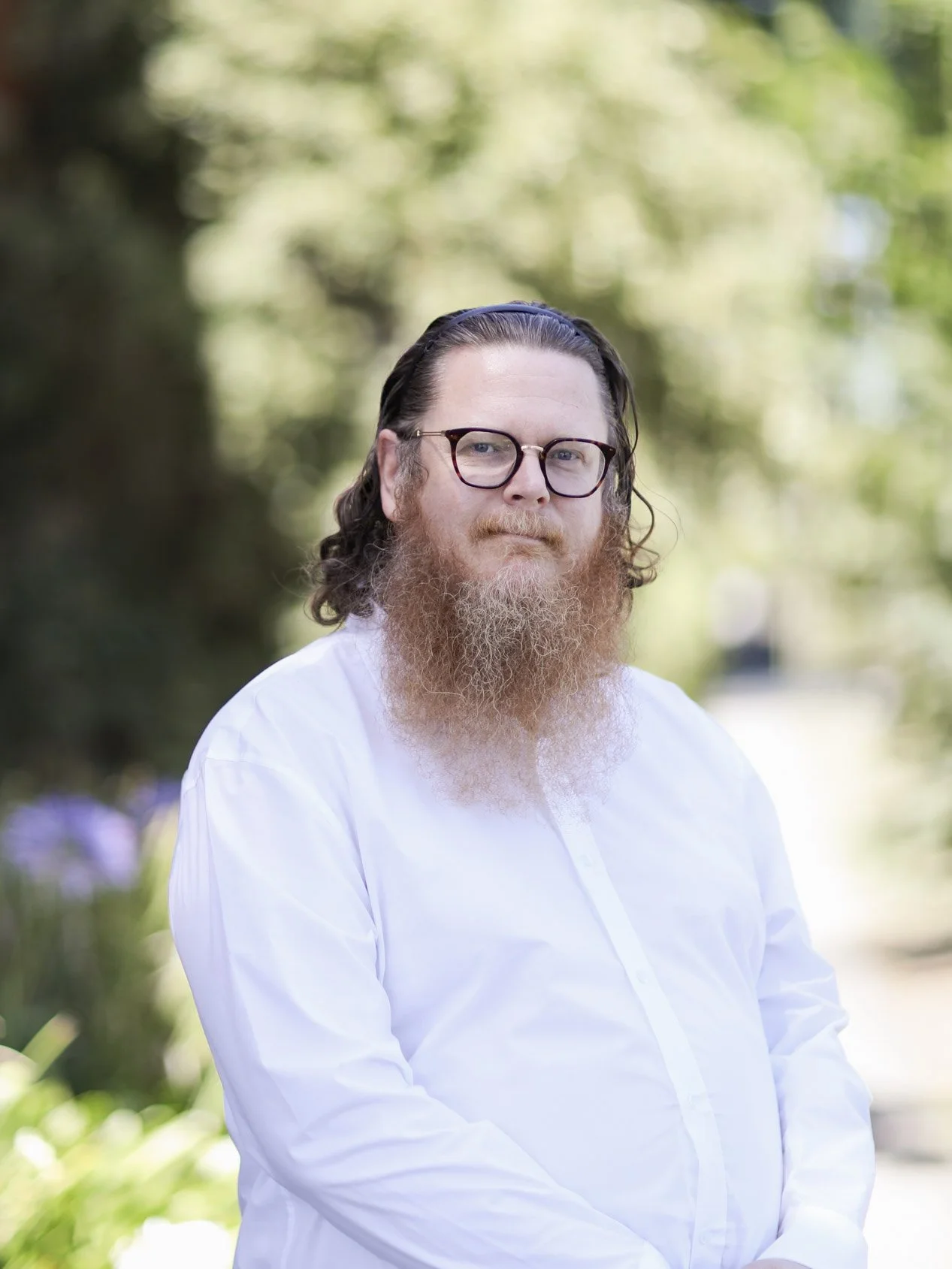 Man with long hair and beard wearing glasses and white shirt outdoors with blurred green trees in background.