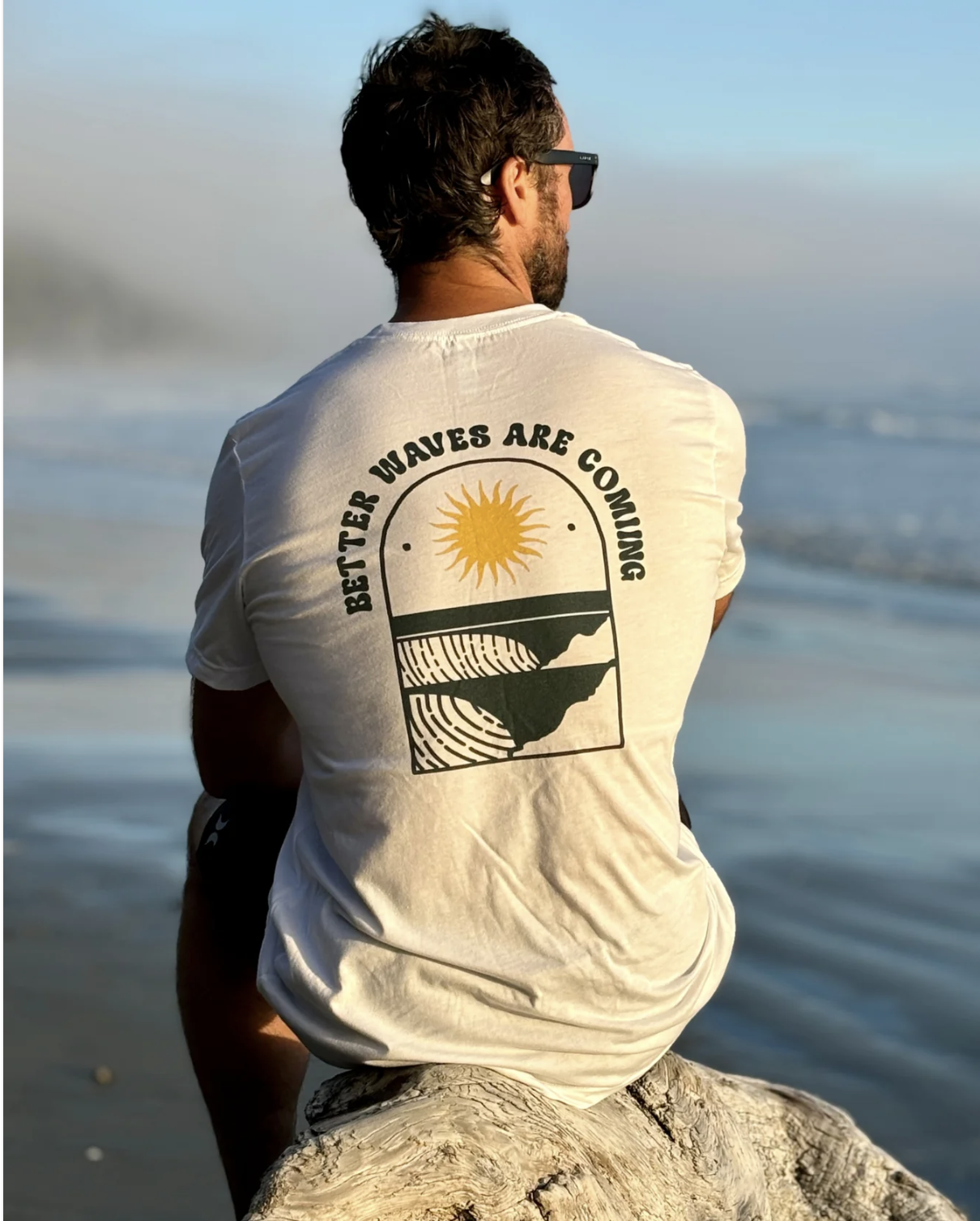 A man sitting on a log at the beach, wearing sunglasses and a beige t-shirt with a design that reads 'Bitter Waves Are Coming' and features a sun, ocean waves, and coastline.