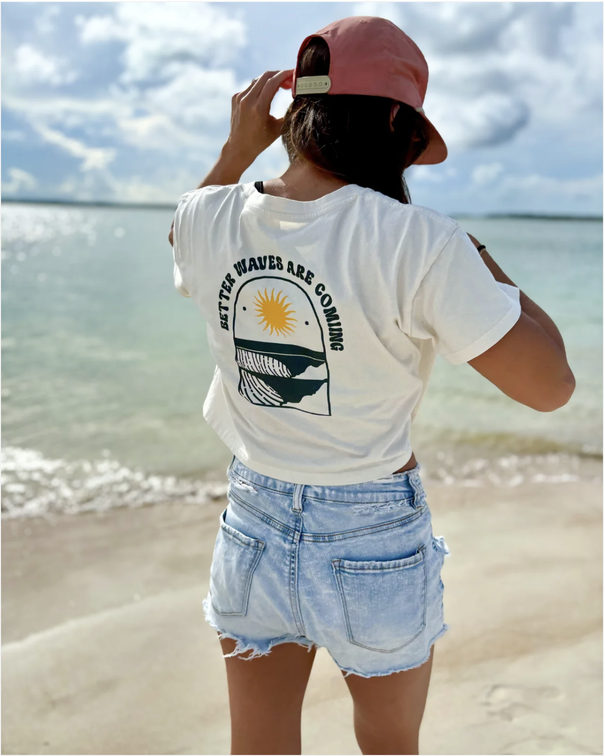 Woman standing on a beach with ocean and cloudy sky in the background. She is wearing a white T-shirt with a graphic and text, light blue denim shorts, and a pink cap.