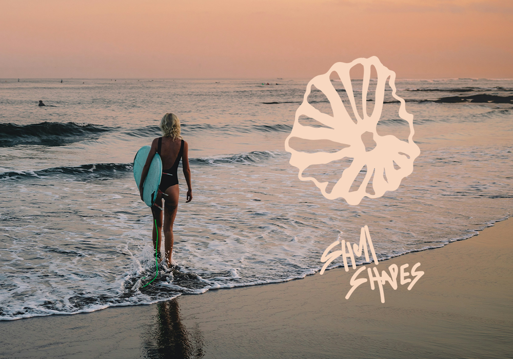 A woman with blond hair carrying a surfboard walks into the ocean at sunset, with a logo and text overlay that read 'Shri Shapes'.}