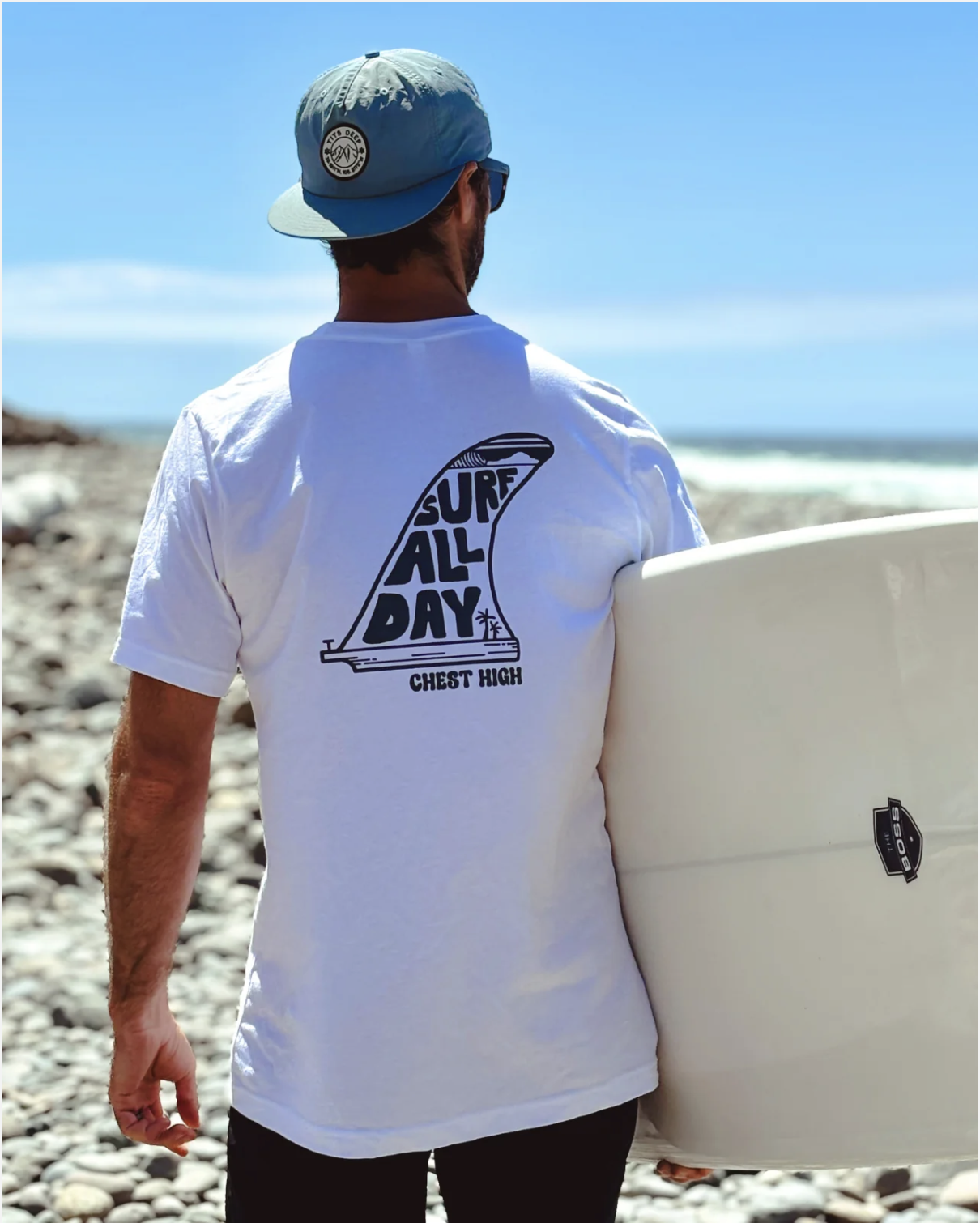 A man holding a surfboard on a rocky beach, wearing a white T-shirt with a surf-related graphic on the back, a light blue cap, and sunglasses, looking toward the ocean under a clear blue sky.