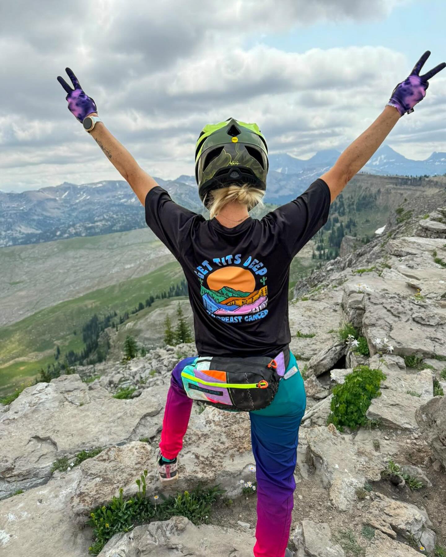Person with blonde hair wearing a black t-shirt, colorful pants, and a helmet, standing on rocky terrain with arms outstretched in a victory pose, overlooking mountain landscape.