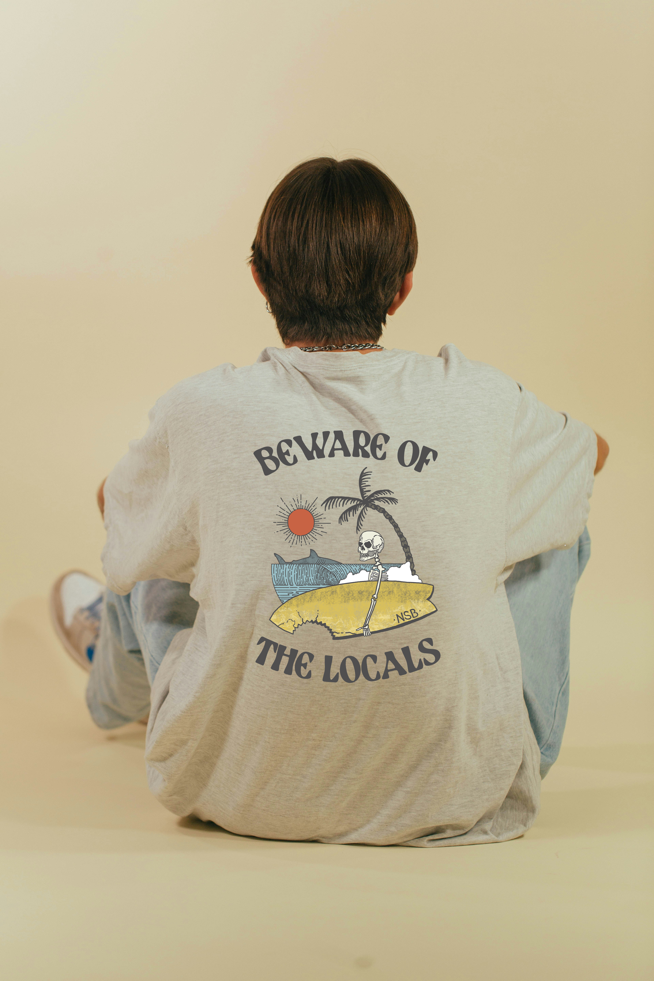Person sitting on the floor with their back facing the camera, wearing a gray t-shirt with a graphic that says 'Beware of the Locals,' depicting a skeleton leaning on a surfboard, with a palm tree, sun, and ocean in the background.